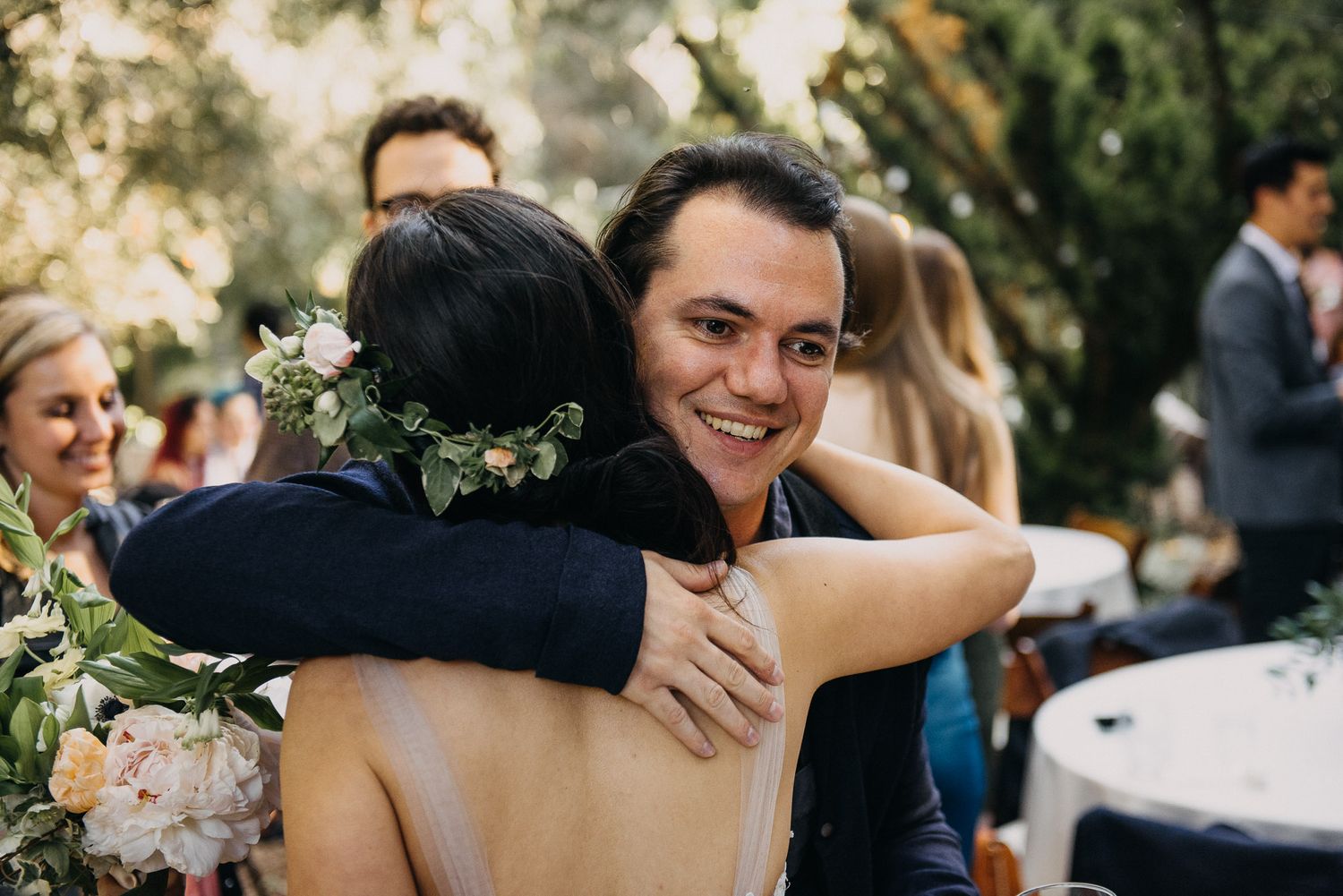 Guests celebrate and embrace during an outdoor wedding reception under natural lighting.