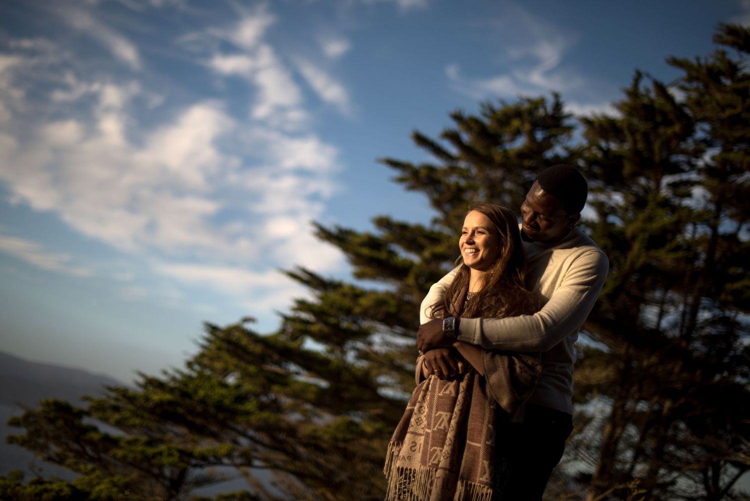 Couple embraces while enjoying sunset views with evergreen trees silhouetted against a dramatic cloudy sky.