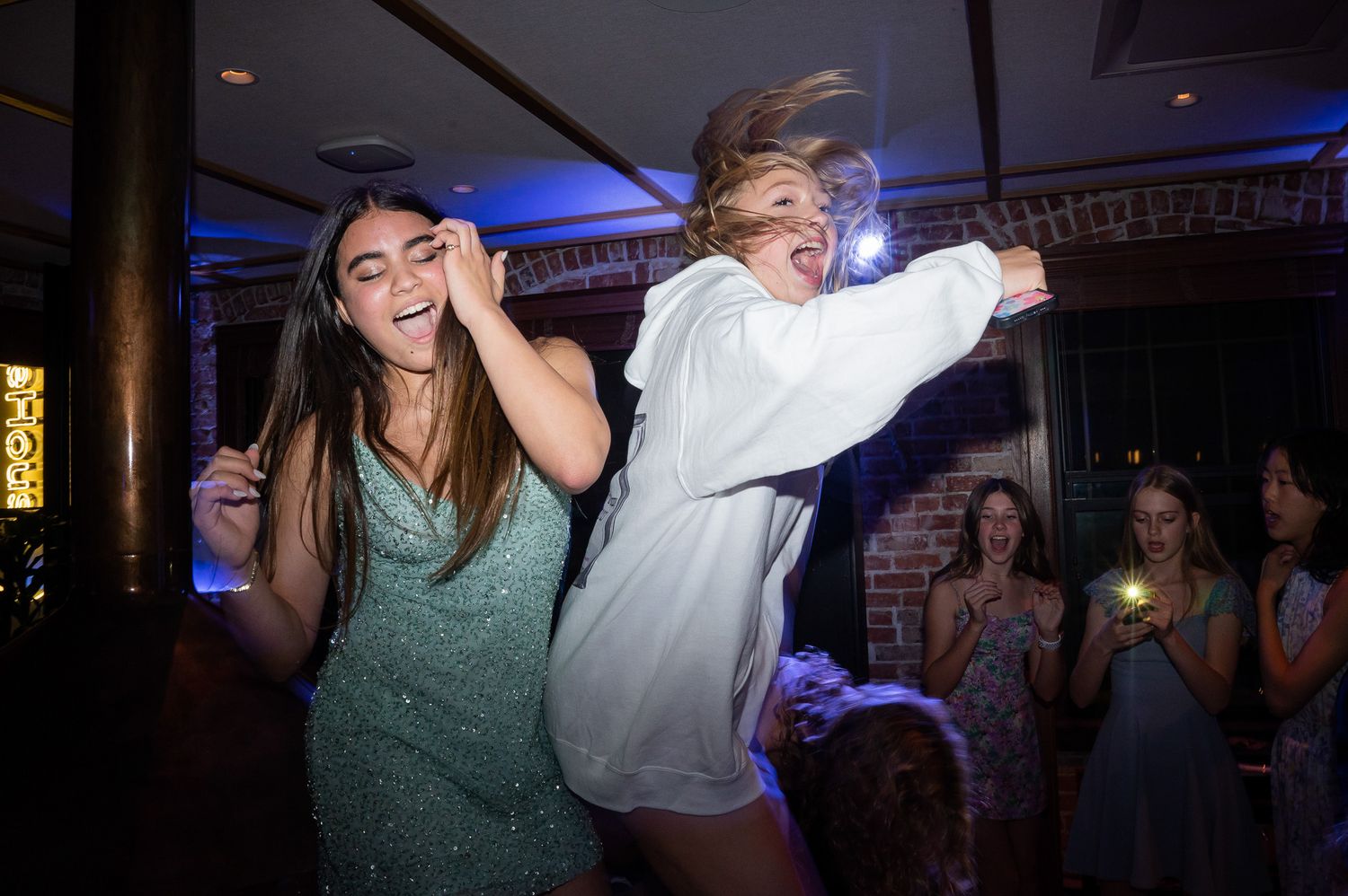 Two people dancing energetically in a dimly lit venue with party guests visible in the background.