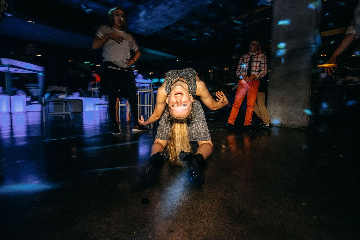 People dance and move energetically on a dark dance floor with dramatic blue lighting and reflective surfaces.