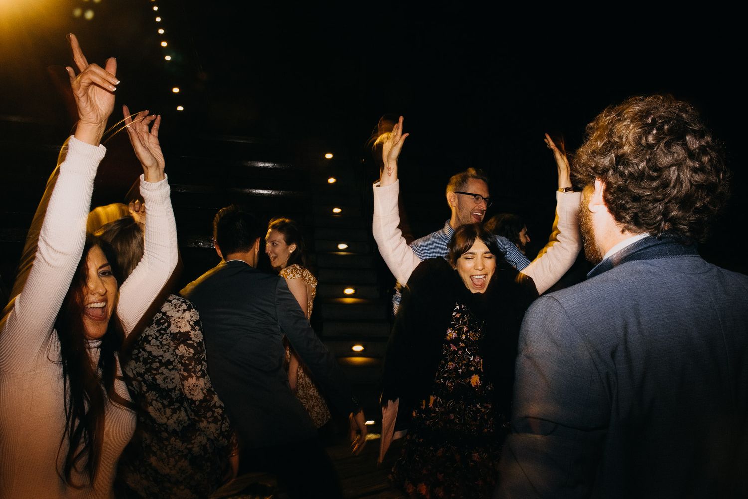 A group of people dancing and celebrating with raised arms at a dimly lit party venue.