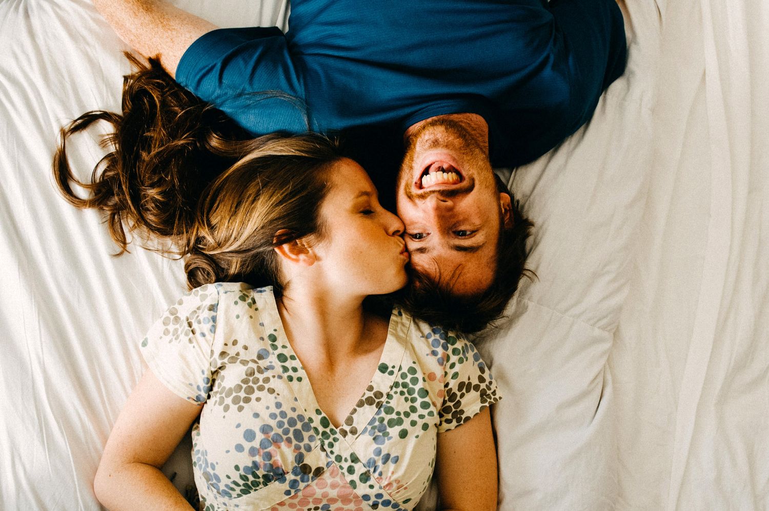 Two people lie together head to head on white sheets, one wearing a floral dress, creating a cheerful close-up portrait.