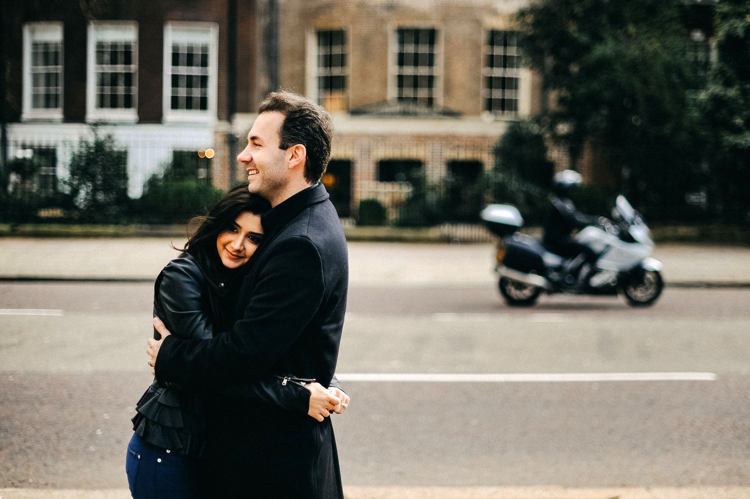 A couple embraces on a city street with a parked motorcycle and historic buildings in the background.