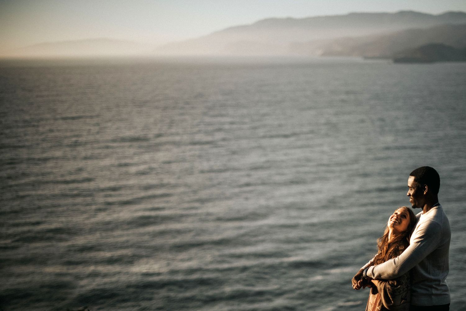 Silhouetted person sits peacefully by the misty ocean cliffs at sunset overlooking the calm sea.