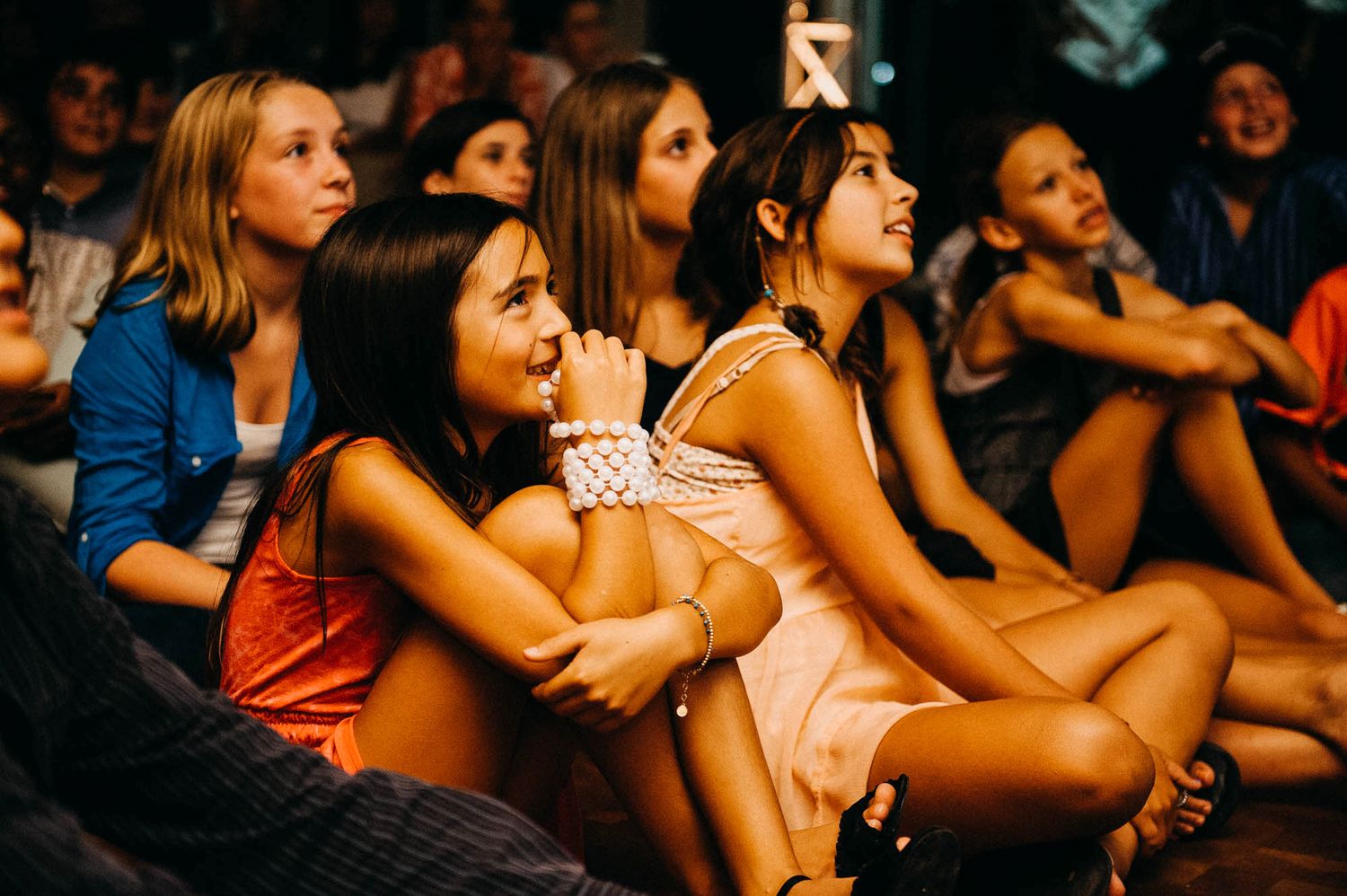 A group of people sit together watching intently at an event or performance in a dimly lit venue.
