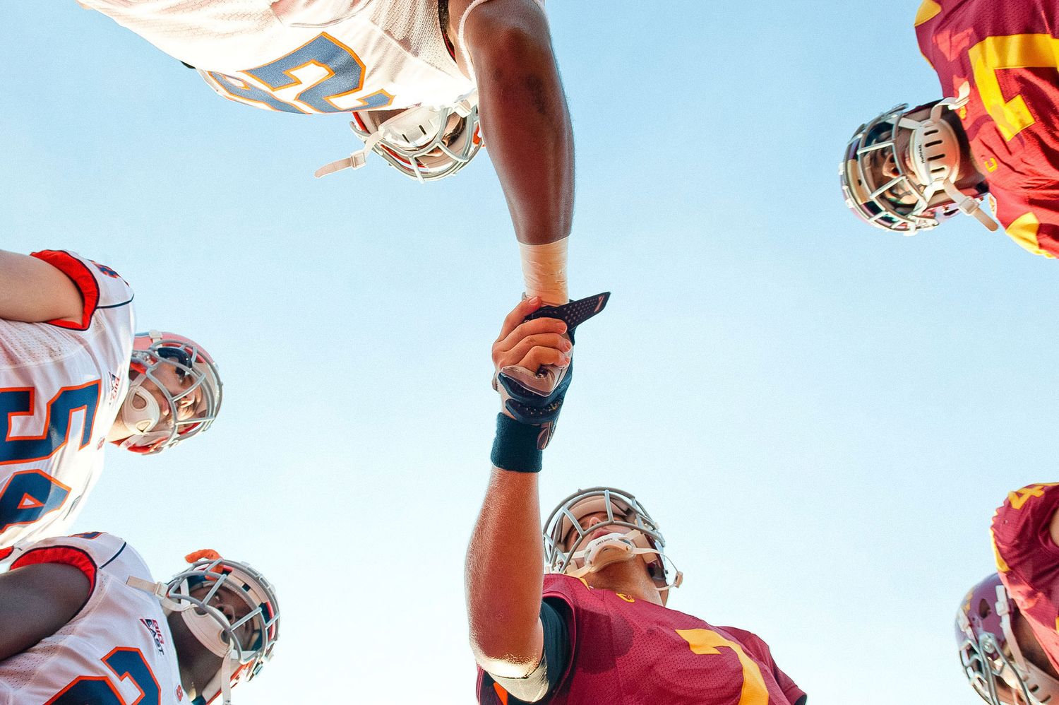 Football players huddle together under blue sky with helmets forming a circle seen from below.