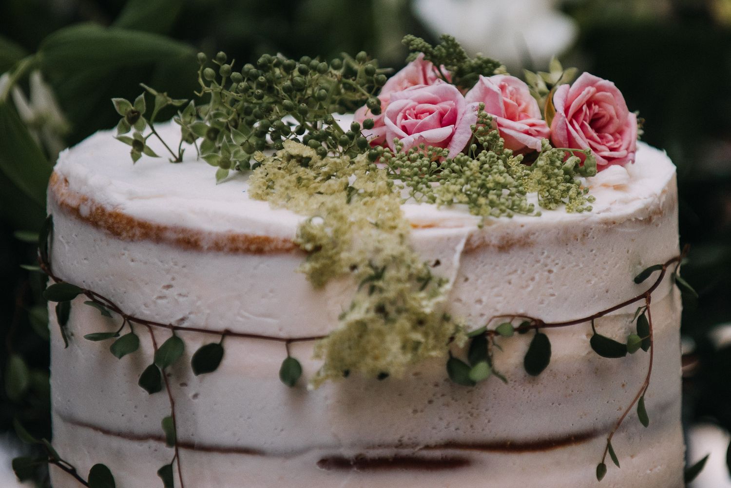 A rustic white wedding cake decorated with delicate pink roses and fresh greenery on a wooden surface.