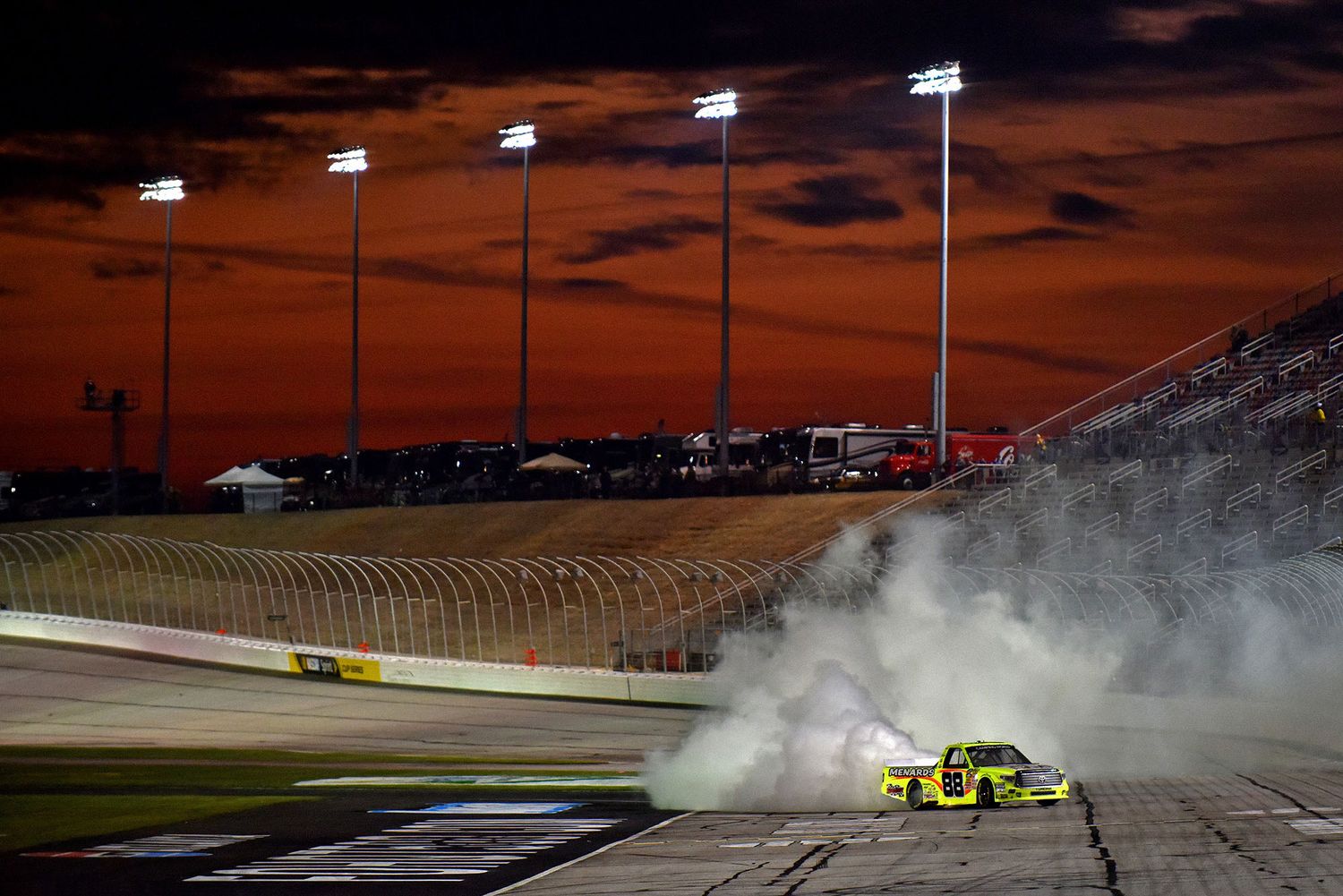 A NASCAR race car does a celebratory burnout under stadium lights against a dramatic orange sunset sky at a speedway.