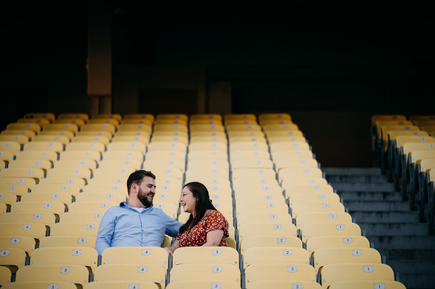 Two people sharing a romantic moment while sitting in empty yellow stadium seats during an engagement photoshoot.