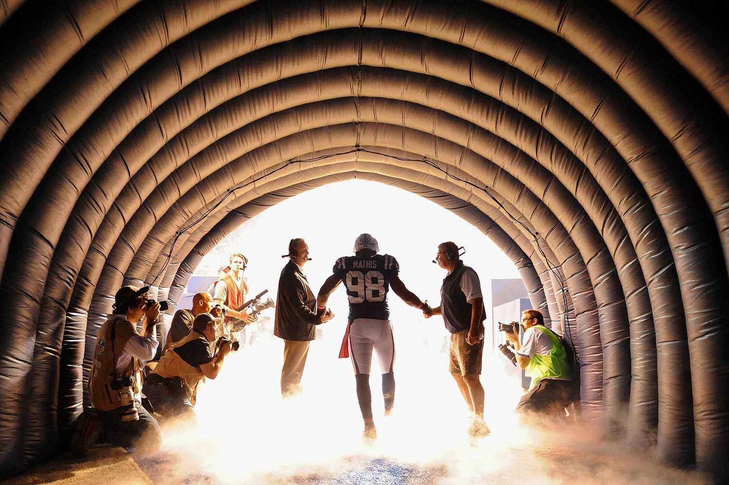 Athletes run through a stadium tunnel silhouetted against bright light while spectators line the sides cheering.