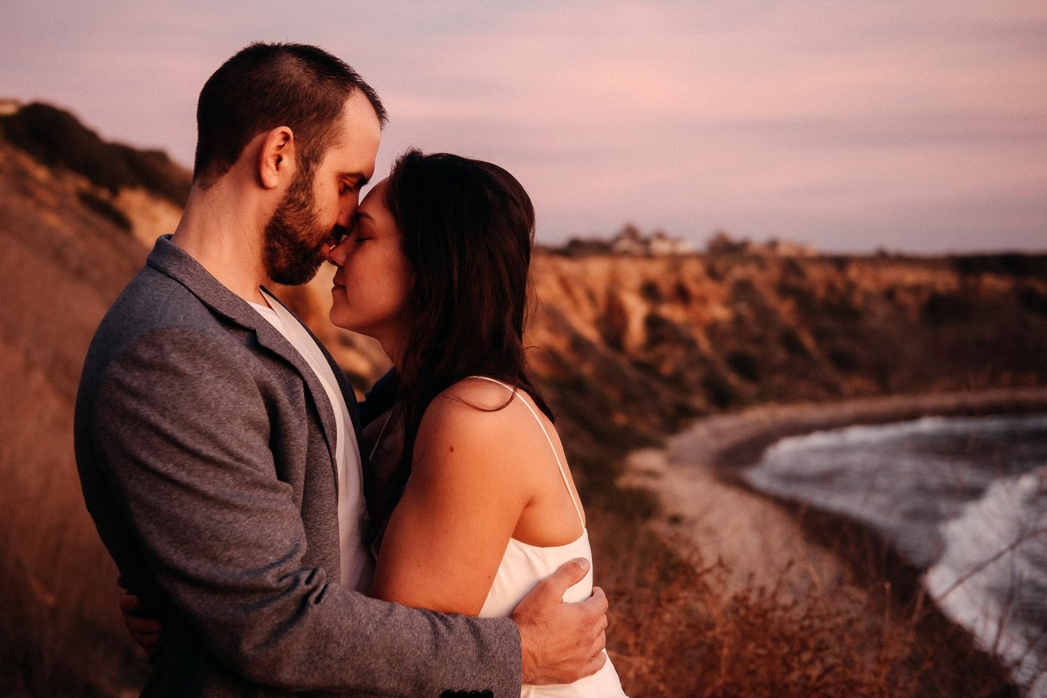 A couple embraces during golden hour on a cliff overlooking a curved beach coastline at sunset.