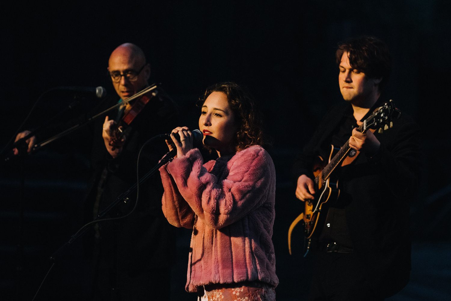 Musicians perform on a dark stage with dramatic lighting during a live concert performance.