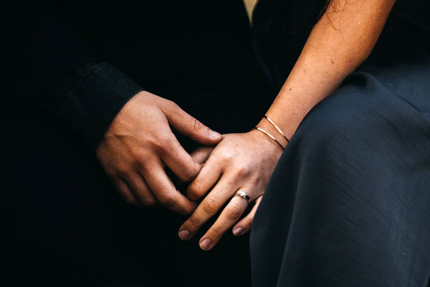 Two hands wearing gold rings and bracelets clasped together against a black background.