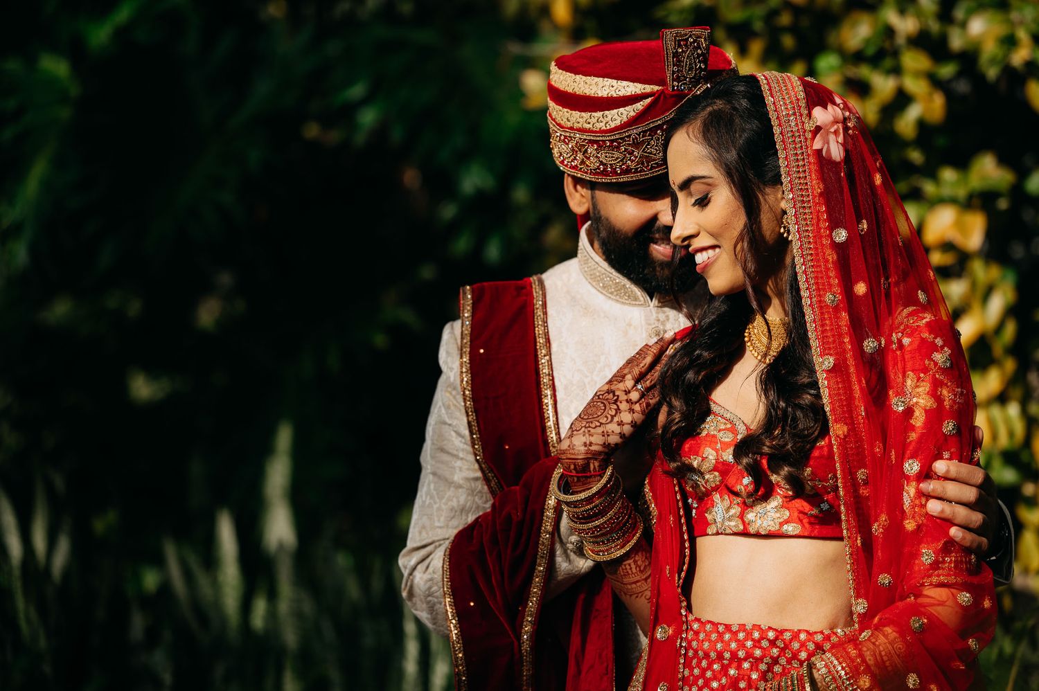 A couple in traditional Indian wedding attire share an intimate moment outdoors in vibrant red and gold outfits.