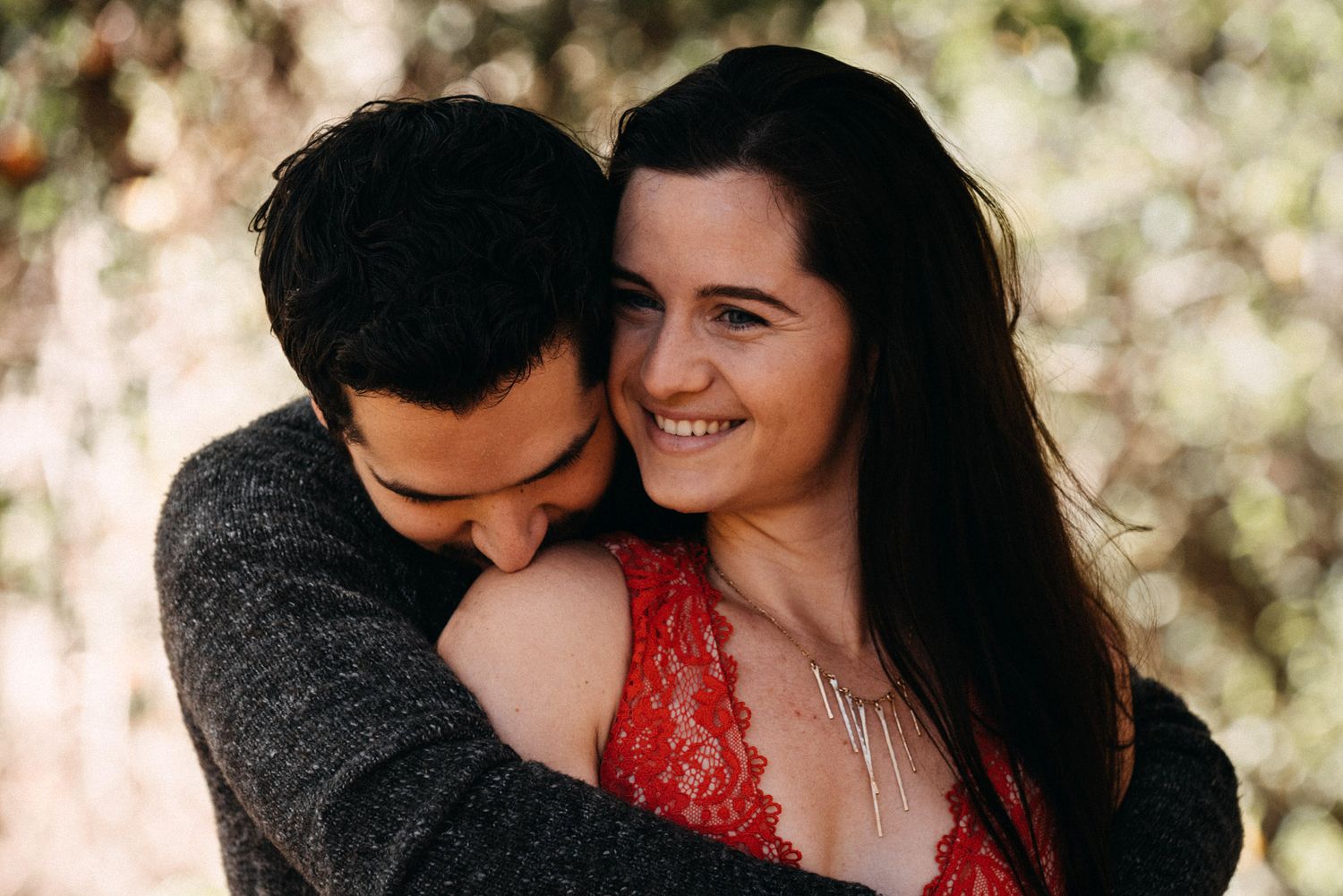 A romantic couple embraces outdoors in warm sunlight while wearing a grey sweater and red lace top.