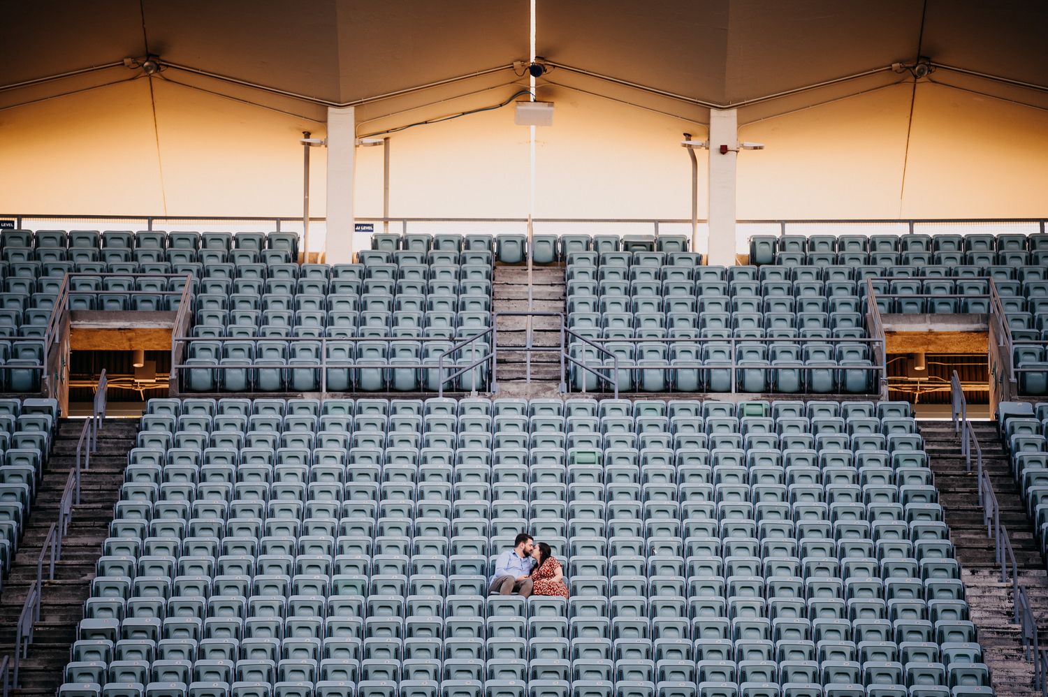 Two spectators sit alone in a vast stadium of empty gray seats under beige ceiling with hanging lights.