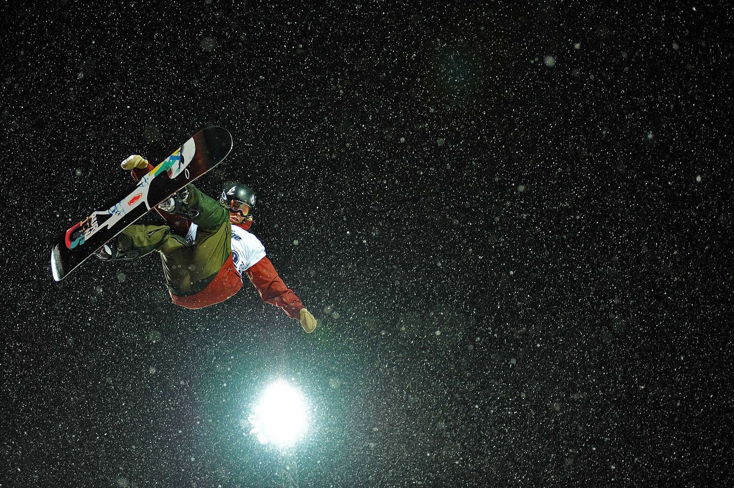 Snowboarder performs aerial trick against night sky with bright moon and stars in background.