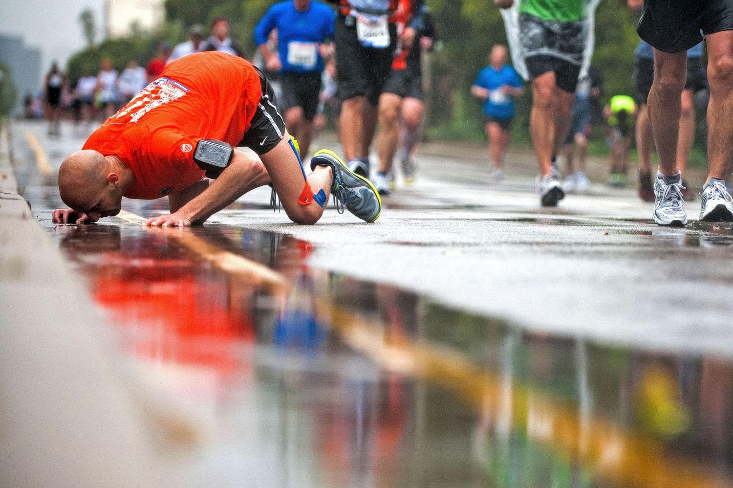 Exhausted runner collapses at finish line of marathon, creating reflection in wet pavement during rainy race day.