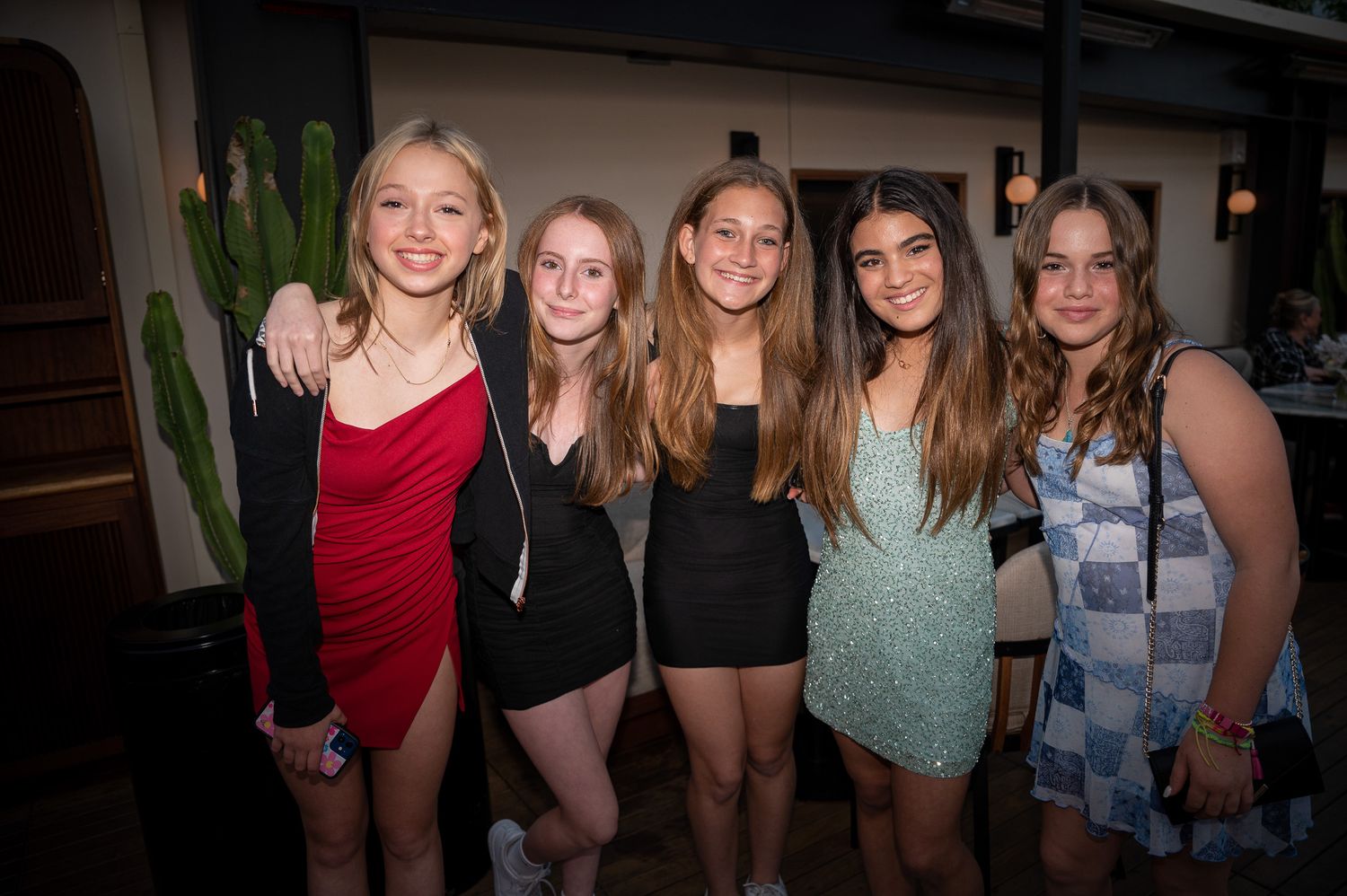 A group of friends pose together at a party wearing cocktail dresses in a dimly lit venue.