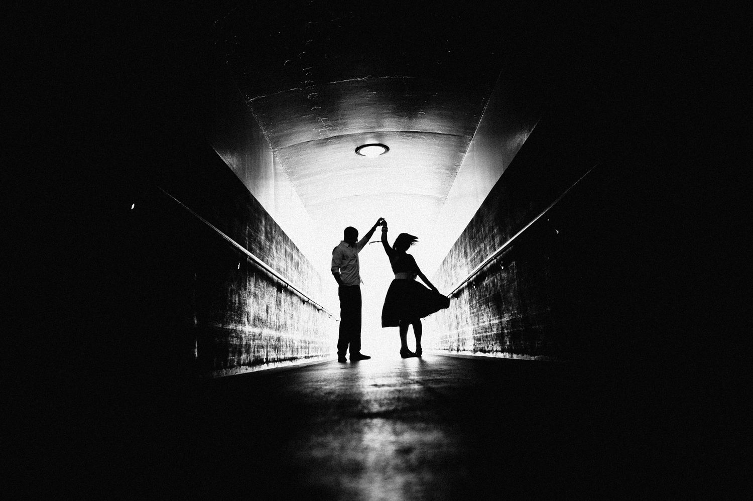 Silhouettes of two people dancing in a tunnel backlit by bright light creating a dramatic black and white scene.