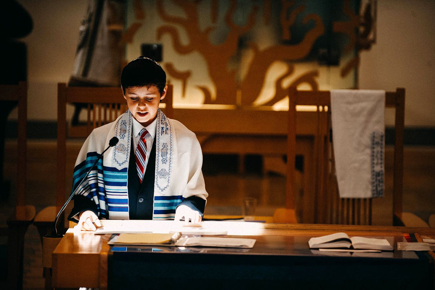 A person wearing a prayer shawl and yarmulke reads from scripture at a wooden lectern during a religious service.
