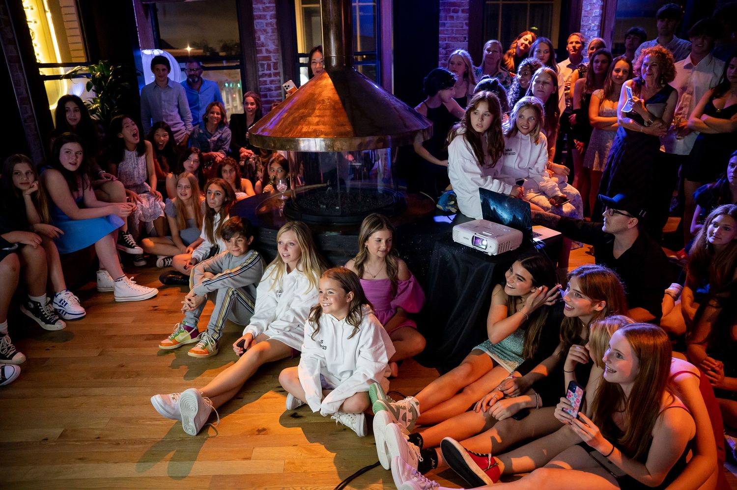 People sitting on a wooden floor watching an indoor performance in a dimly lit venue with purple lighting.