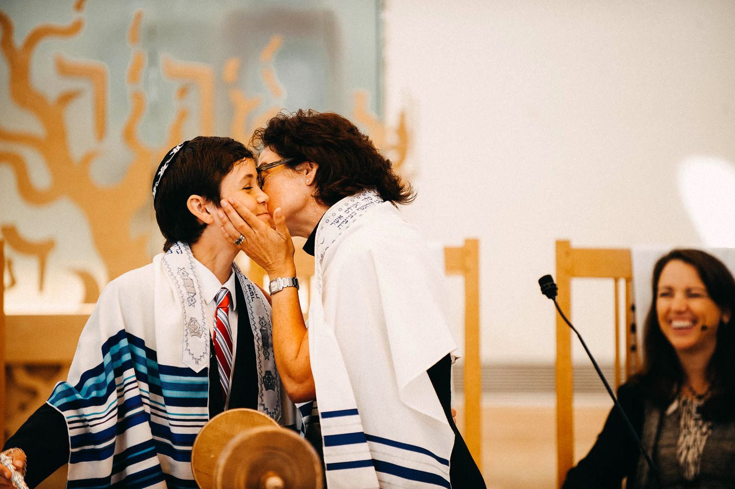 Two people in prayer shawls share a joyful moment during a Jewish celebration while someone smiles in the background.