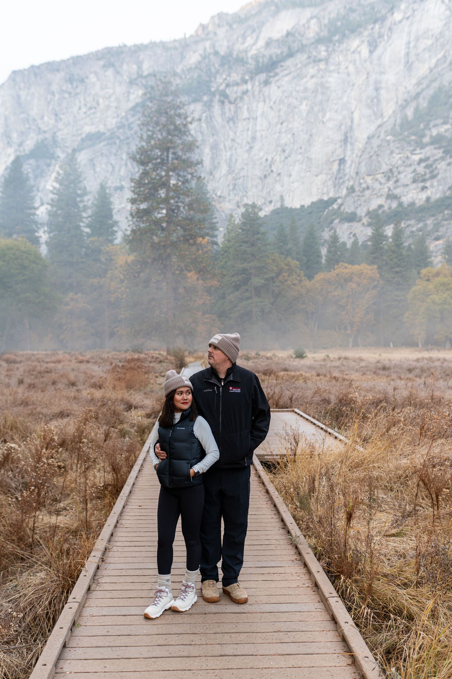 A couple sits together on a wooden boardwalk with majestic mountain cliffs and autumn meadow views.