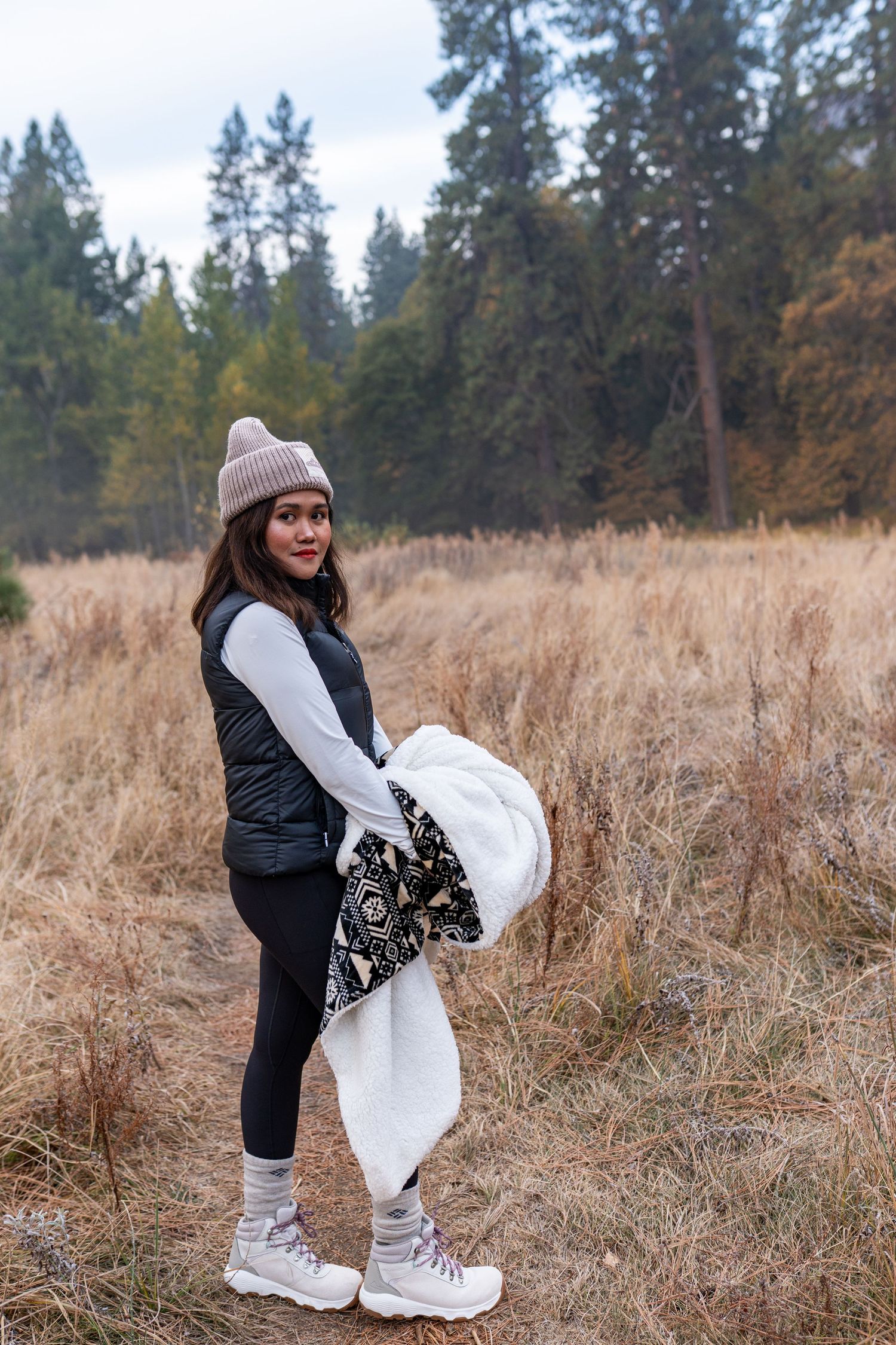 Someone in winter attire poses playfully in a field of tall grass with evergreen trees in the background.