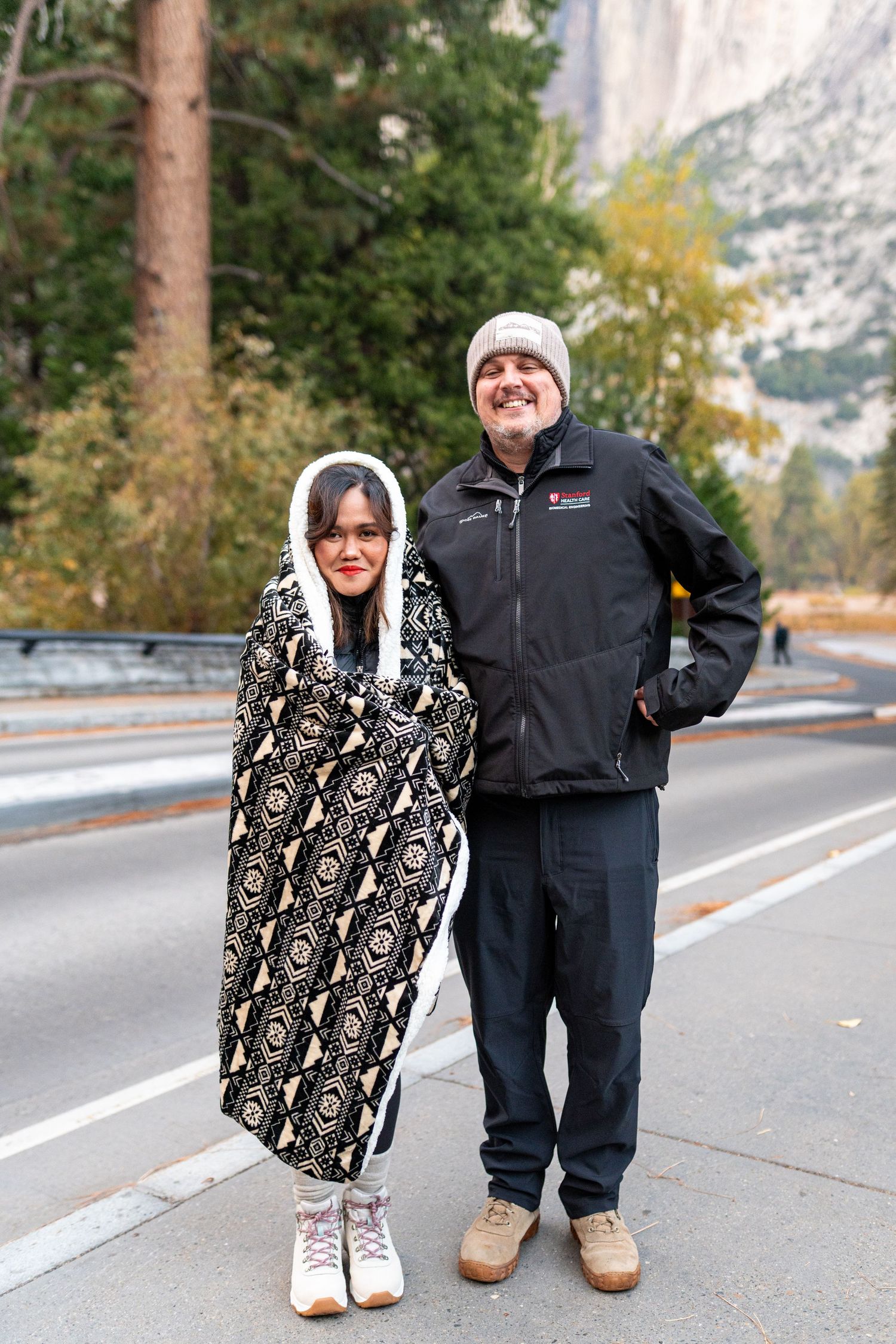 Two people posing together on a mountain road with scenic cliffs in the background.