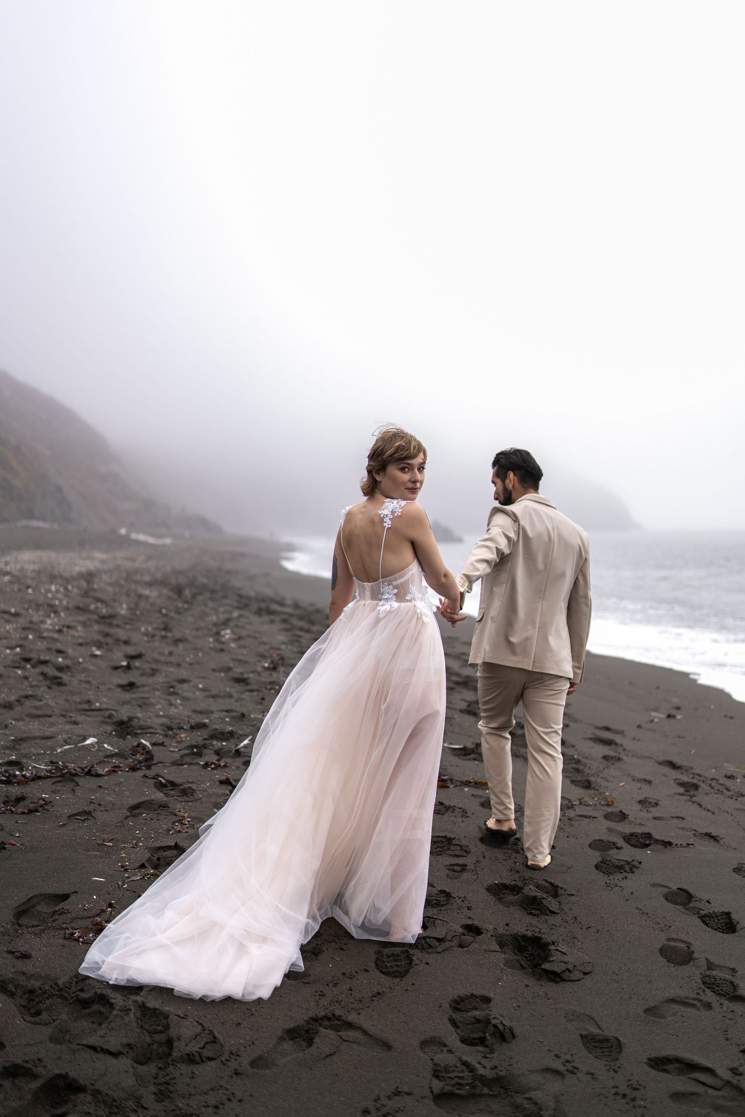 Bride in white gown and groom in beige suit hold hands on foggy black sand beach with misty ocean backdrop.