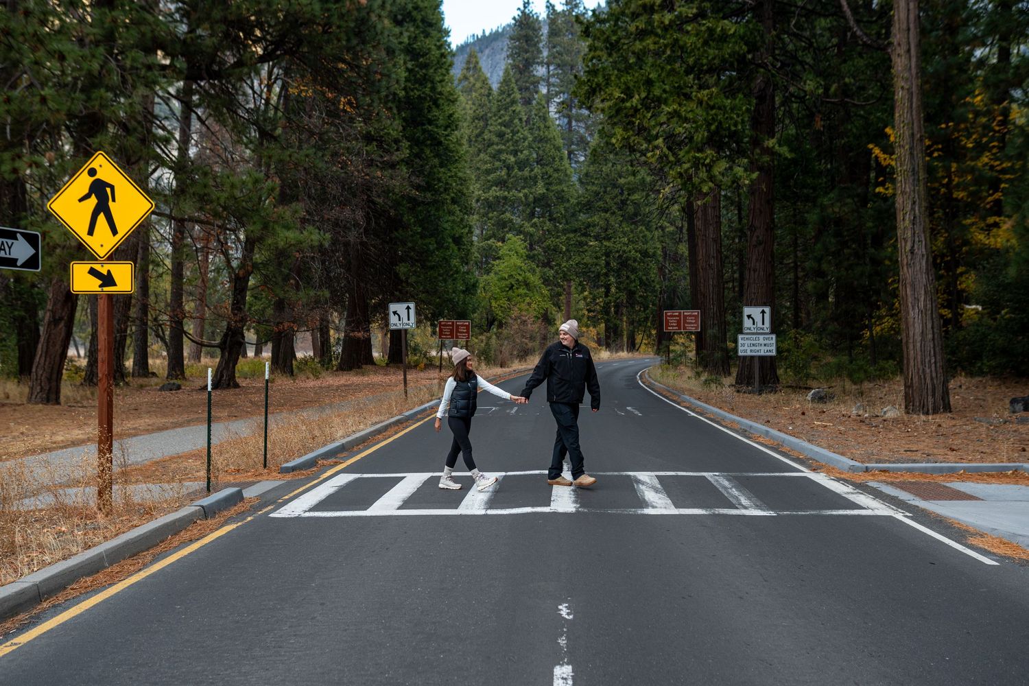 Two people walking across a crosswalk on a forest road with pedestrian crossing signs.