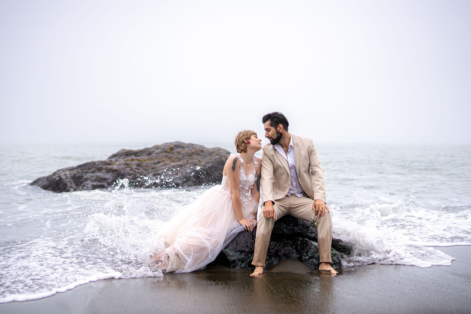 Romantic couple in wedding attire laying in ocean waves at beach during sunset photoshoot.