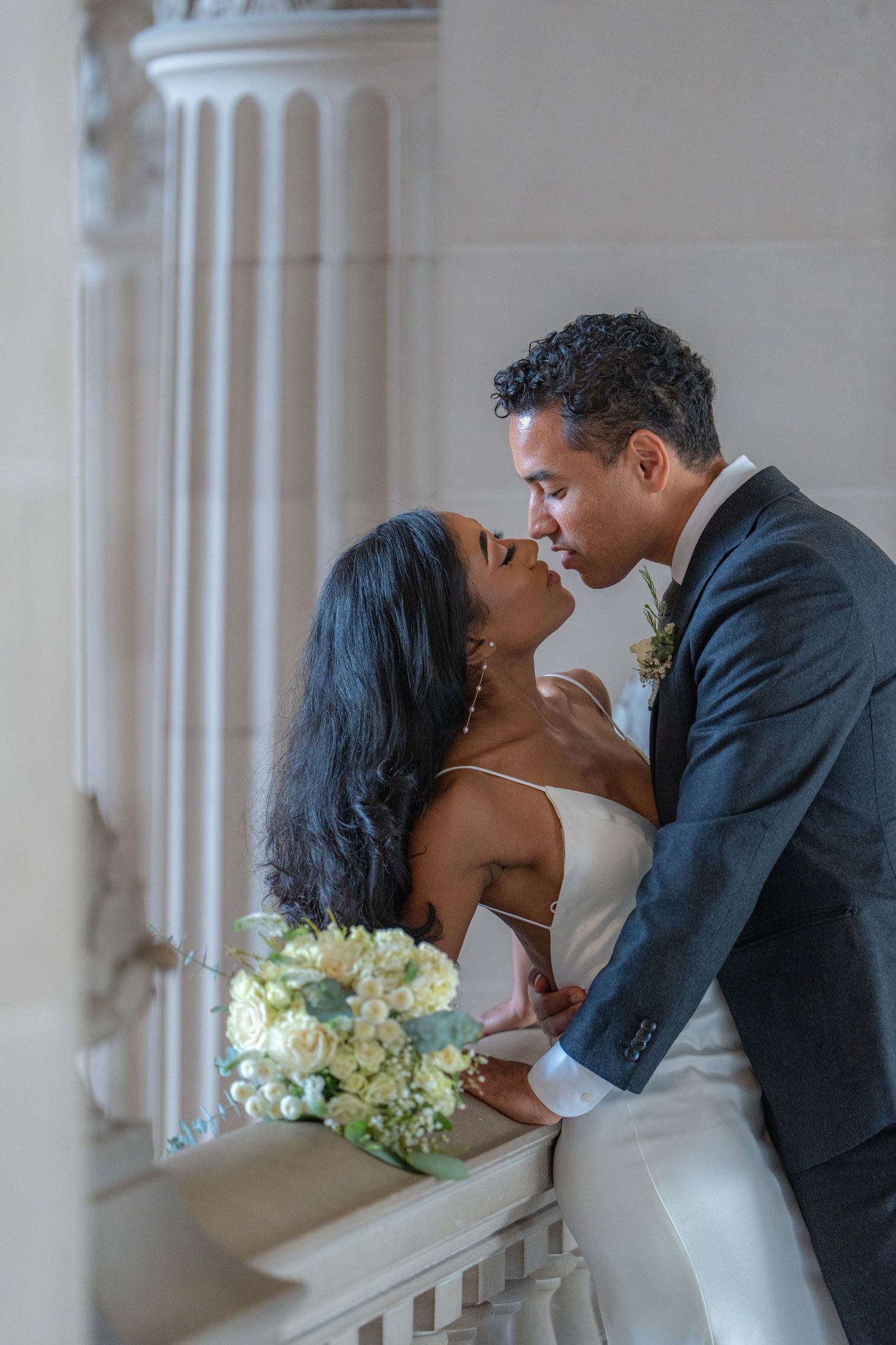 Romantic wedding couple shares intimate moment in elegant white hallway with floral bouquet.