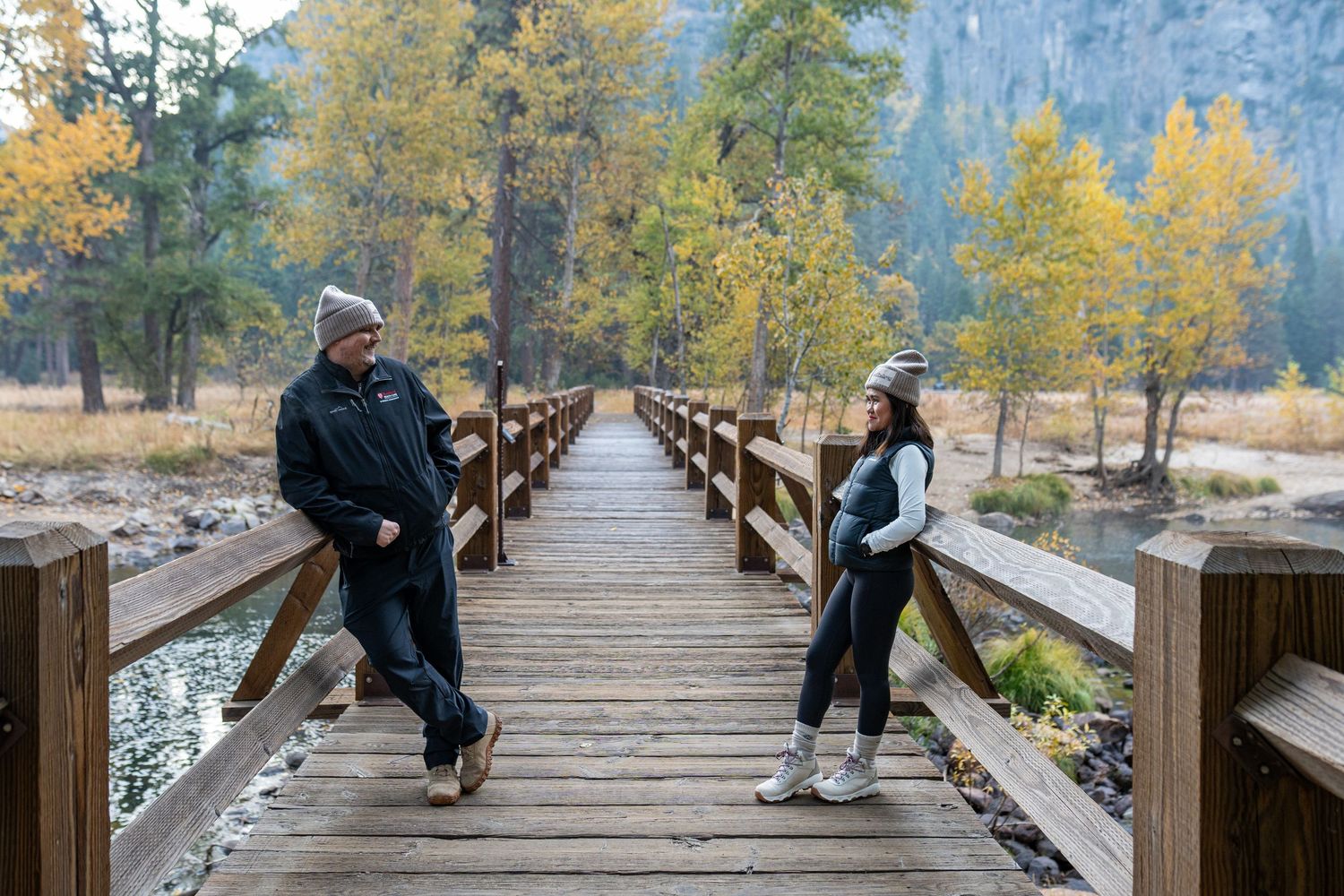 Two people sit and pose together on a long wooden boardwalk with mountains and fall colors in view.