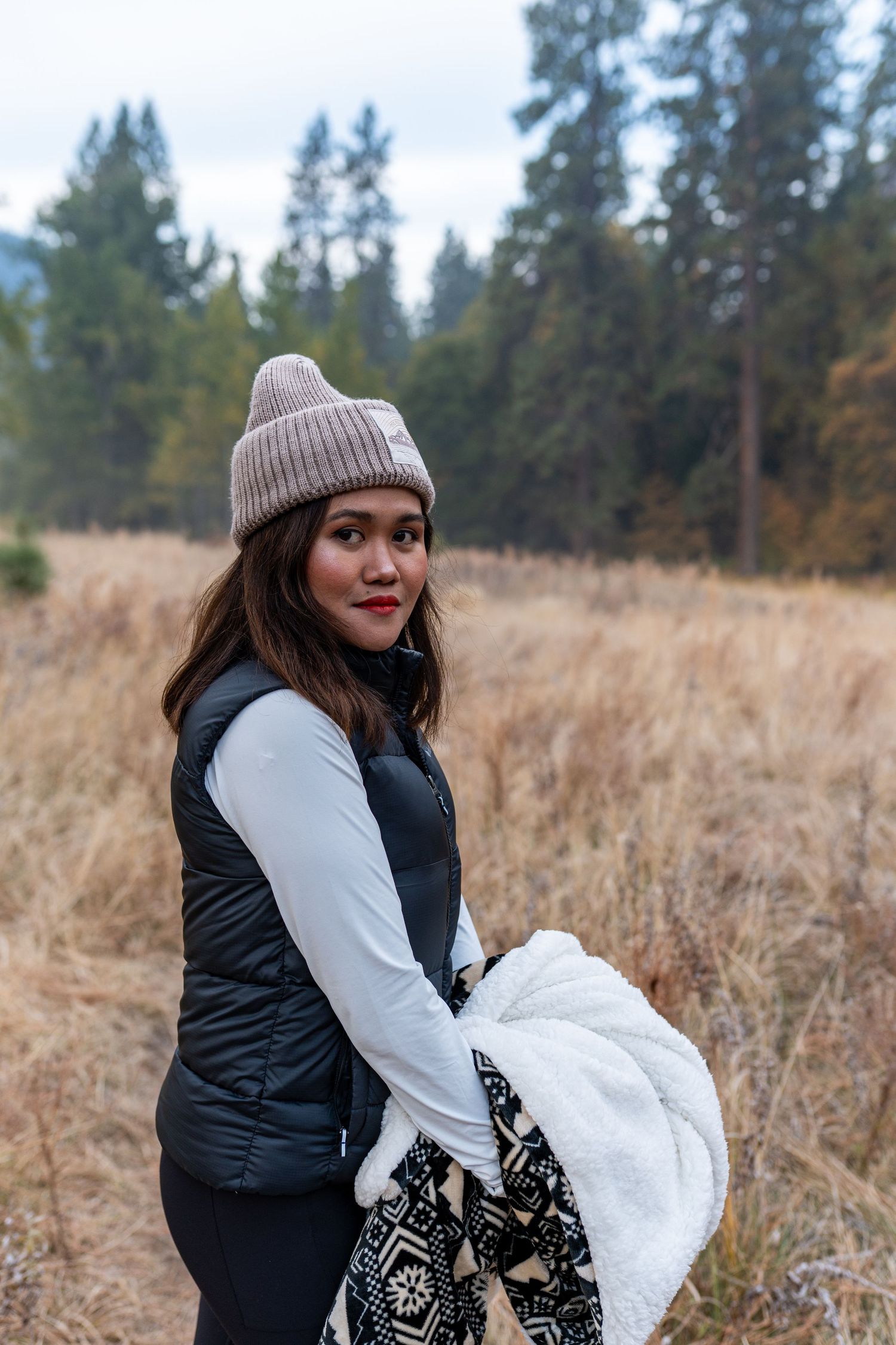 A person in a beanie and vest smiles while posing in an autumn field with pine trees behind.