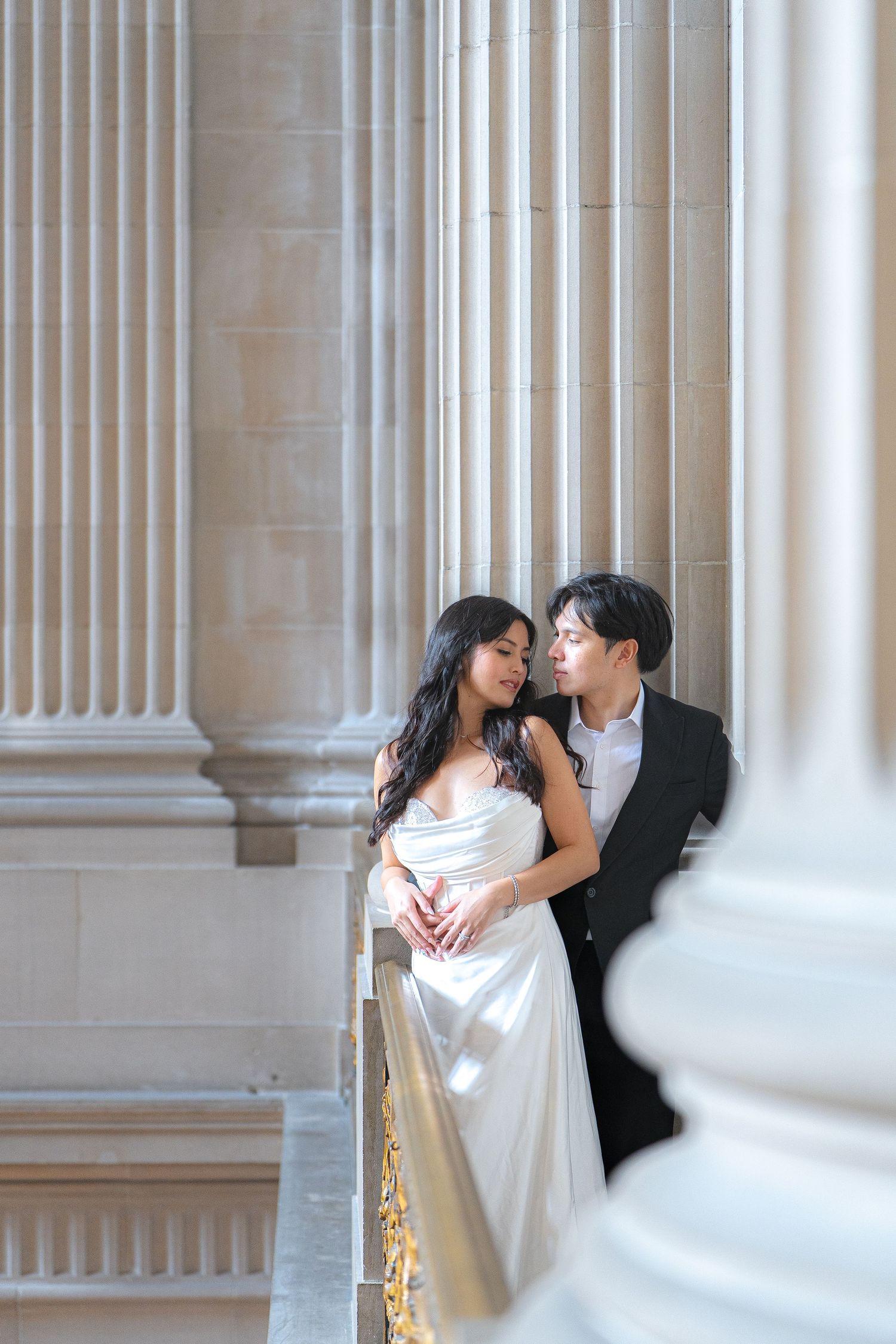 A couple in wedding attire share an intimate moment between elegant white columns in a classic architectural setting.