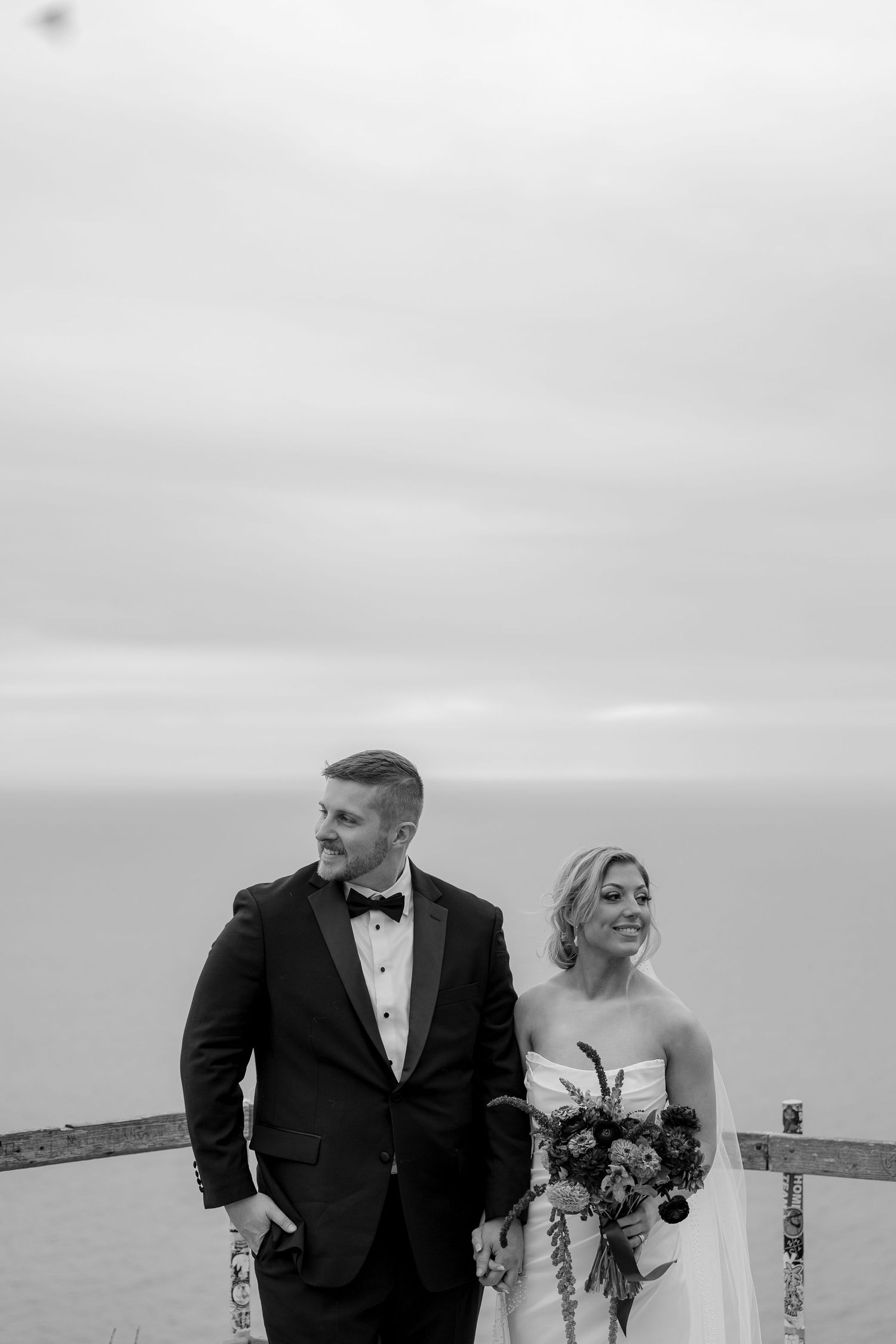 A bride and groom share a joyful moment together on their wedding day by the water in black and white photography.