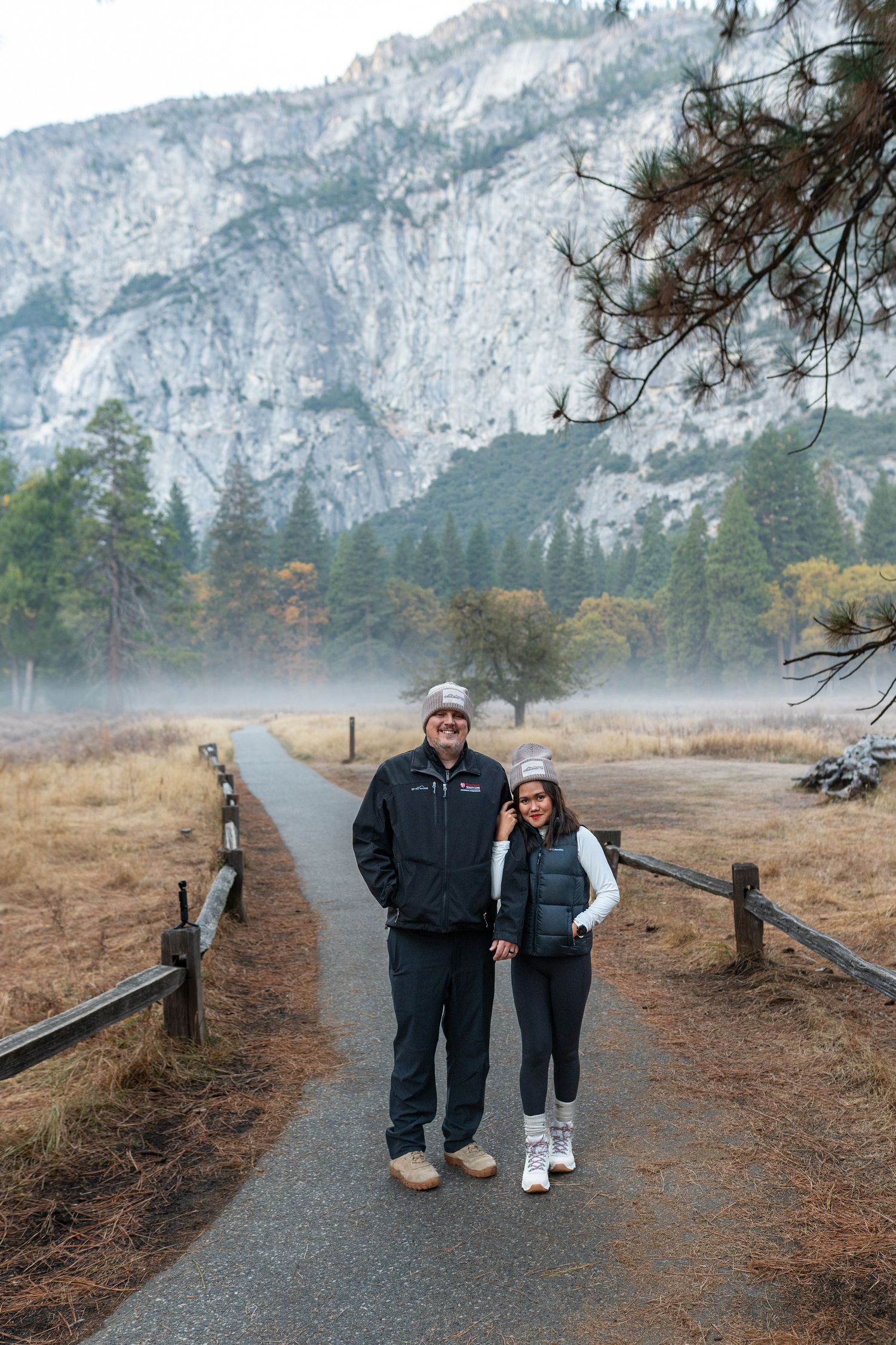 Two people walk along a gravel path with towering granite cliffs and autumn trees in the background.