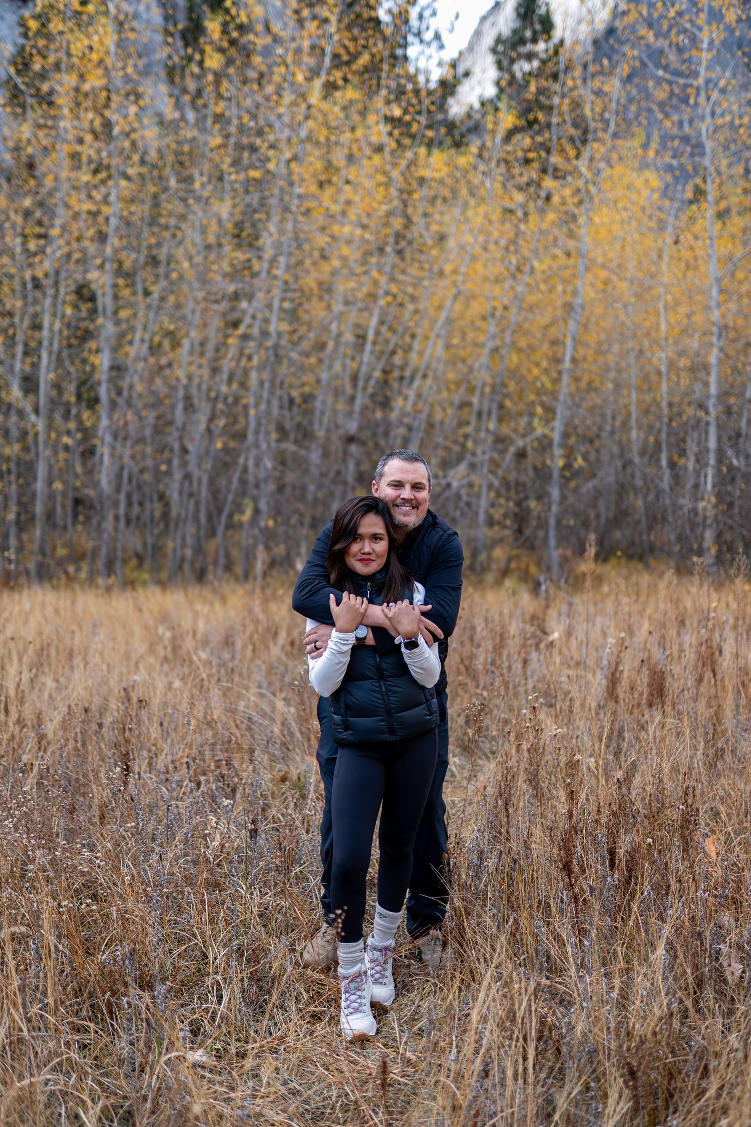 A playful moment captured between two people in a field of tall grass with autumn trees behind.
