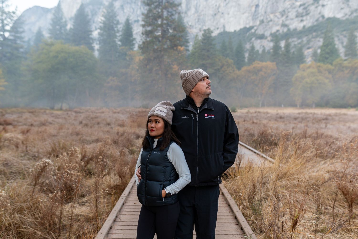 A couple embraces while walking on an elevated wooden path through a mountain meadow.