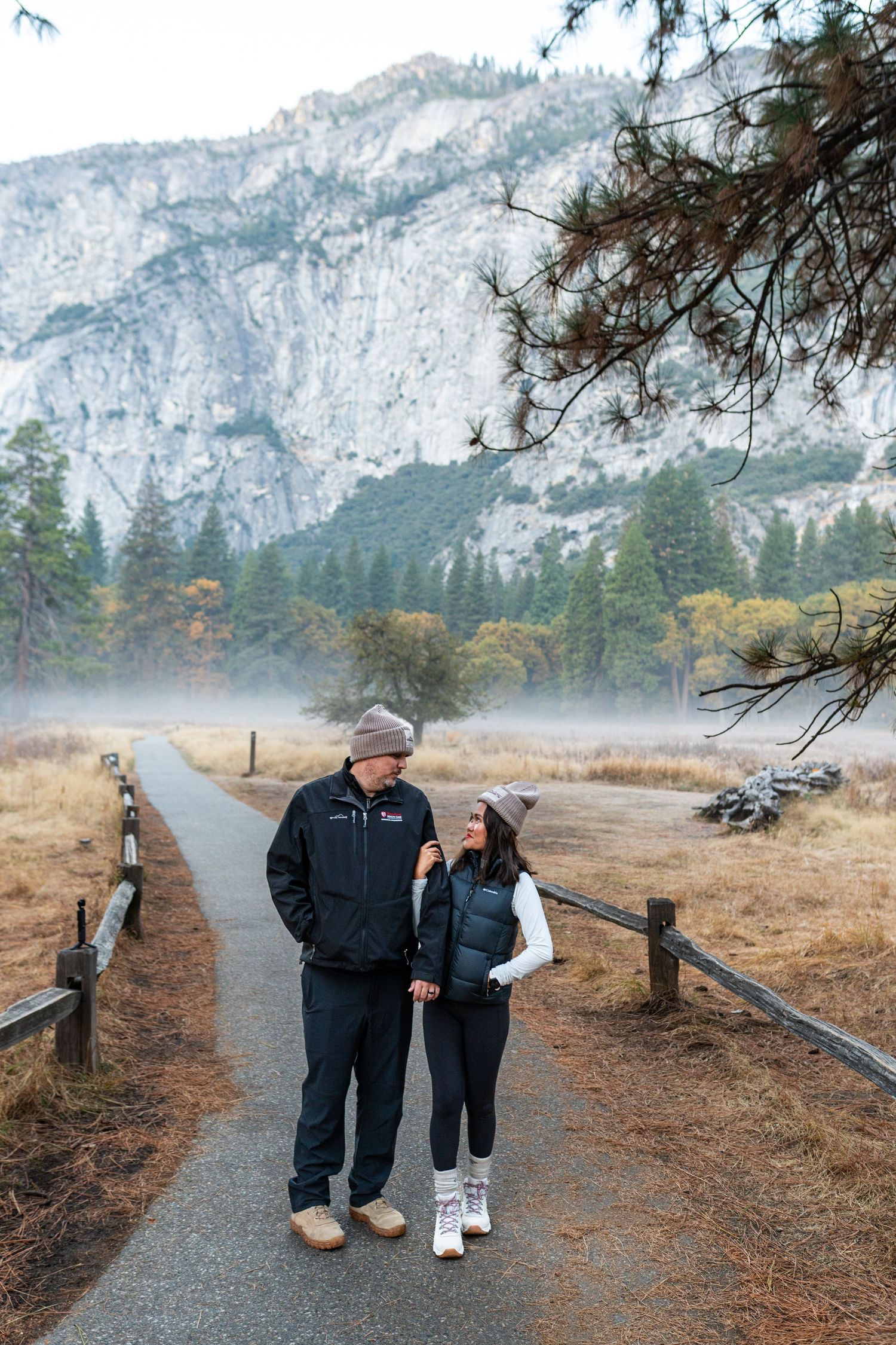 A couple strolls down a scenic mountain trail surrounded by misty cliffs and fall foliage.