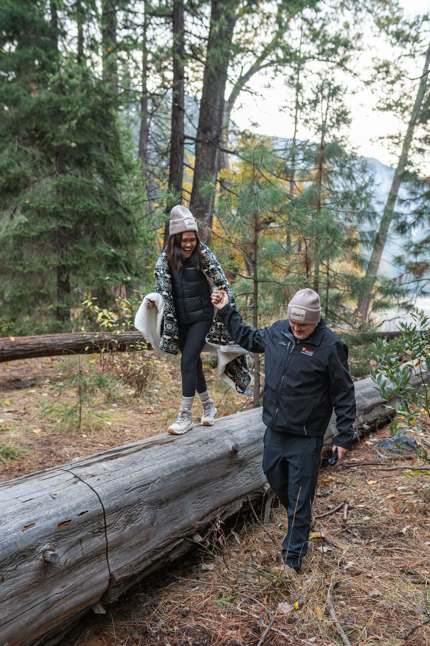 People balancing on fallen logs in a forested area with mountains visible in the distance.