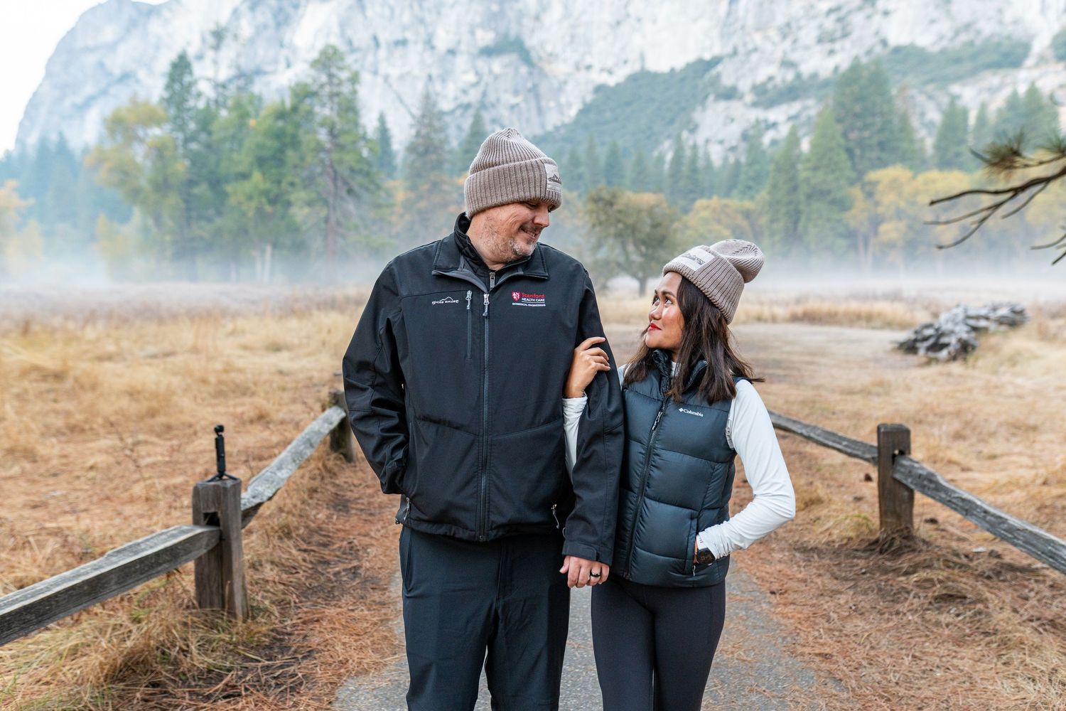 Two people walk hand in hand on a trail with dramatic mountain cliffs rising in the background.