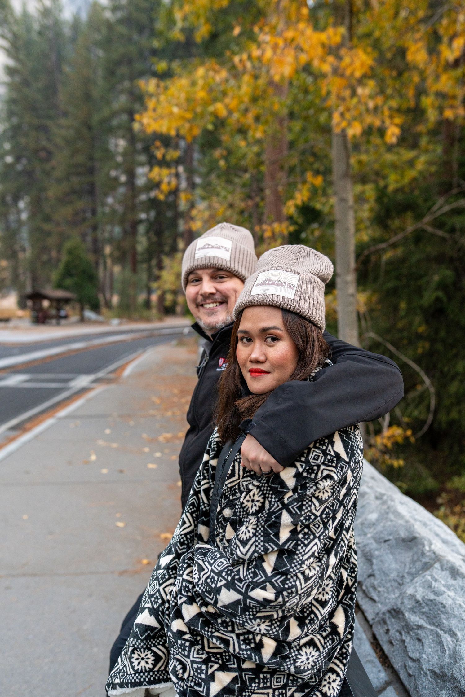 A couple in matching winter outfits pose playfully against a backdrop of fall foliage and a mountain road.
