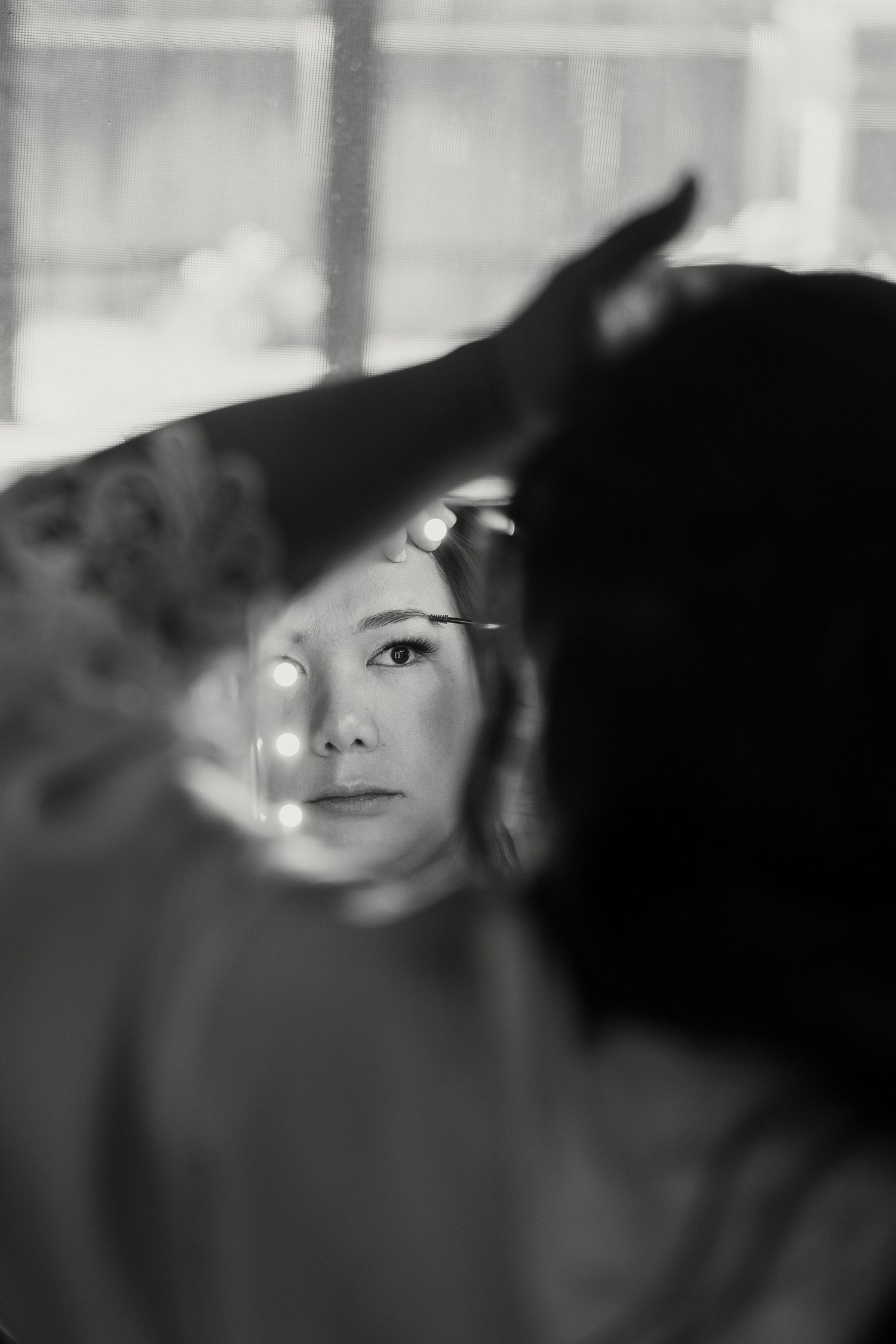 Black and white photo of a woman getting makeup applied, her reflection visible with bokeh lights in background.