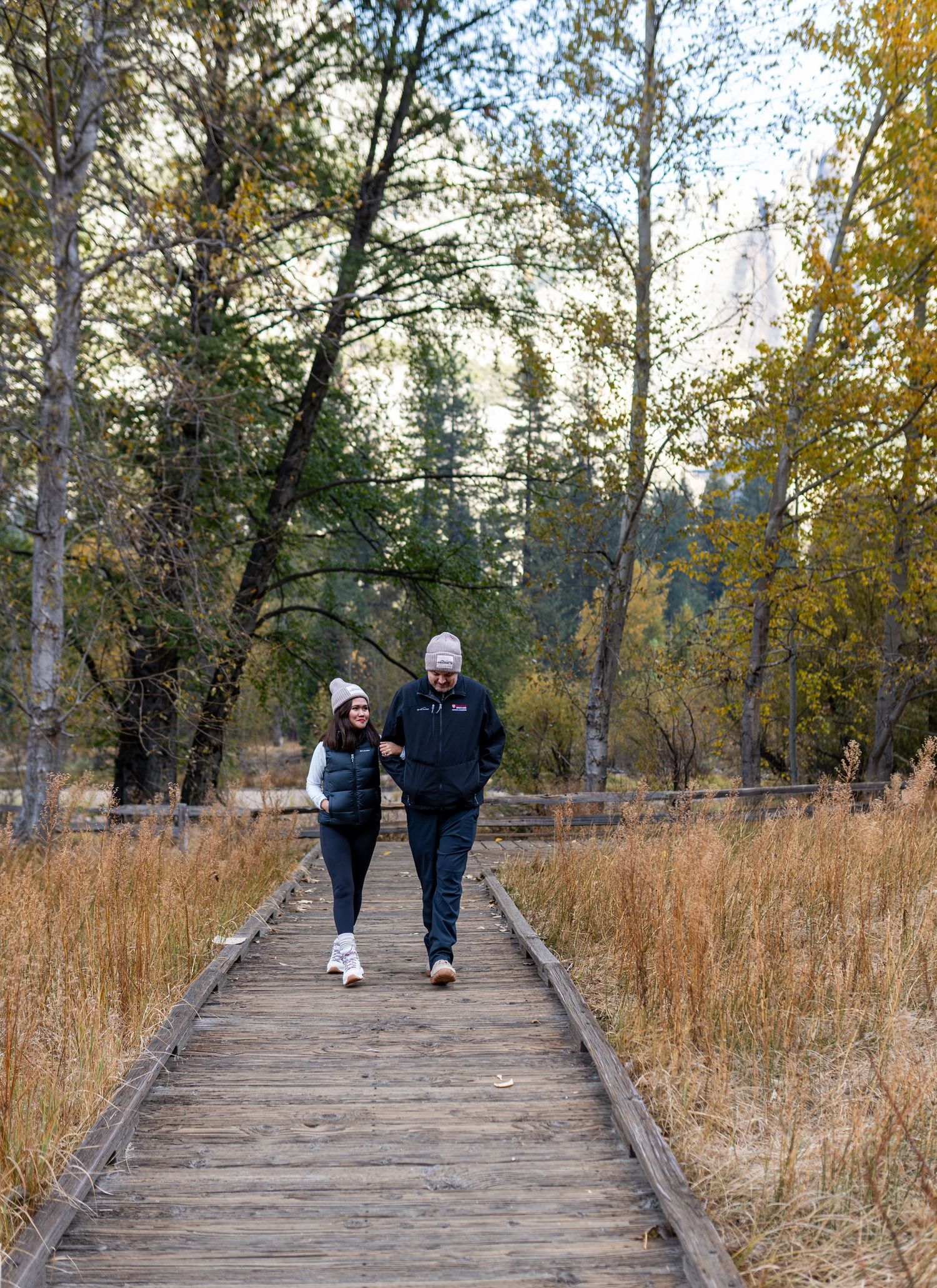 Two people walking together on a wooden boardwalk through autumn grasslands.