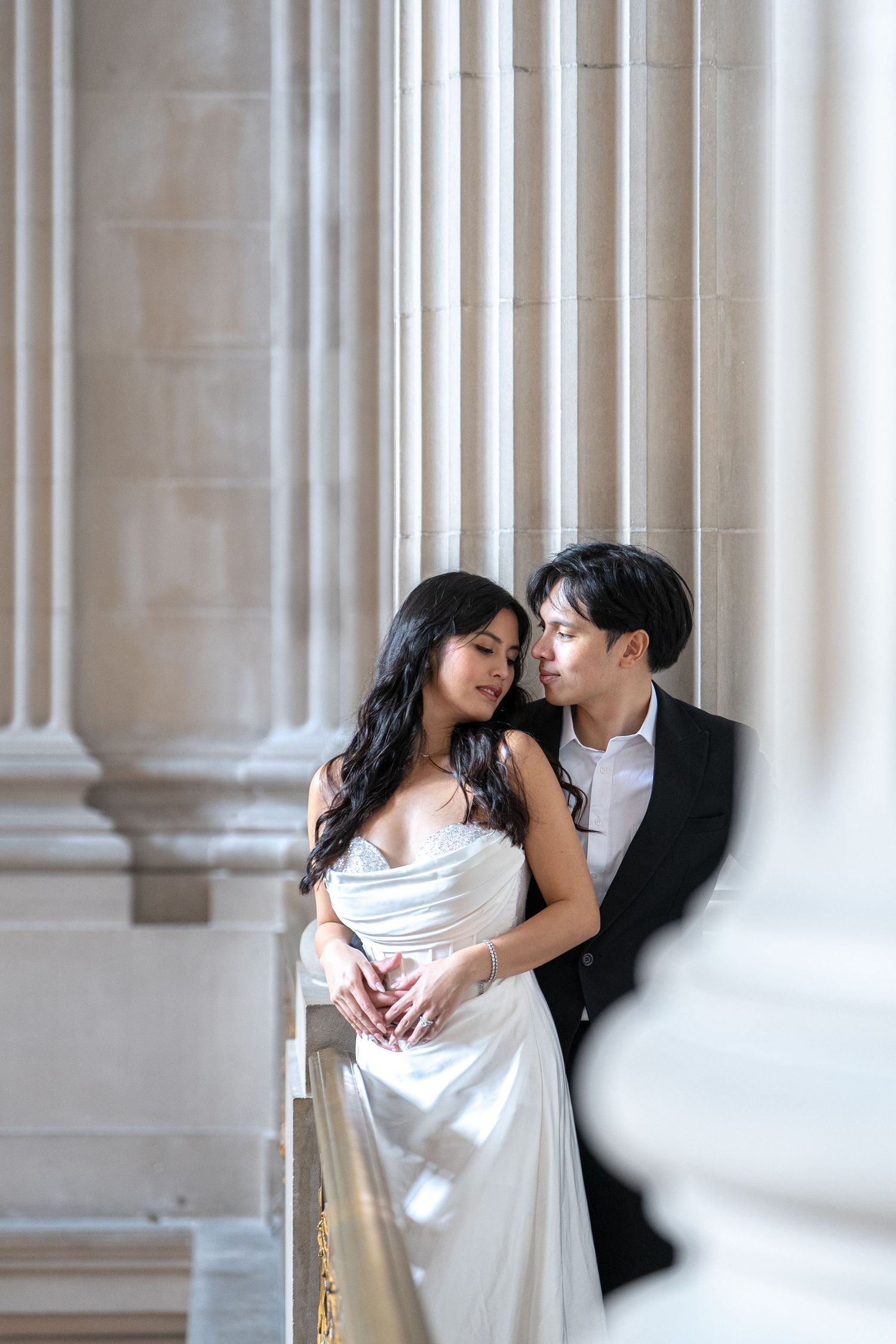 A couple in wedding attire embraces romantically between classical stone columns in an elegant architectural setting.
