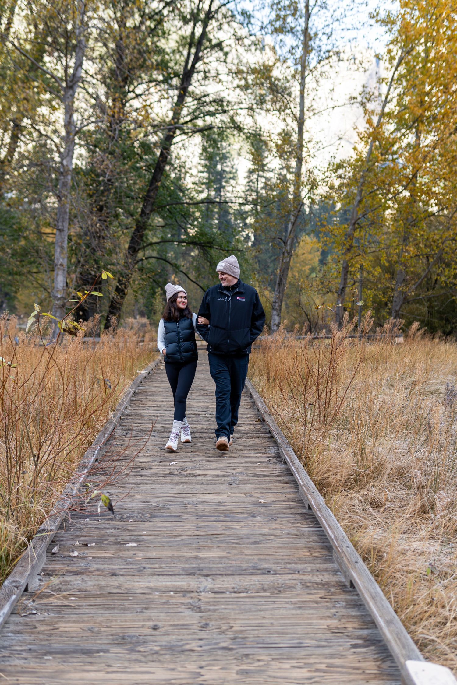 Two people strolling along a wooden boardwalk through golden autumn grasses.