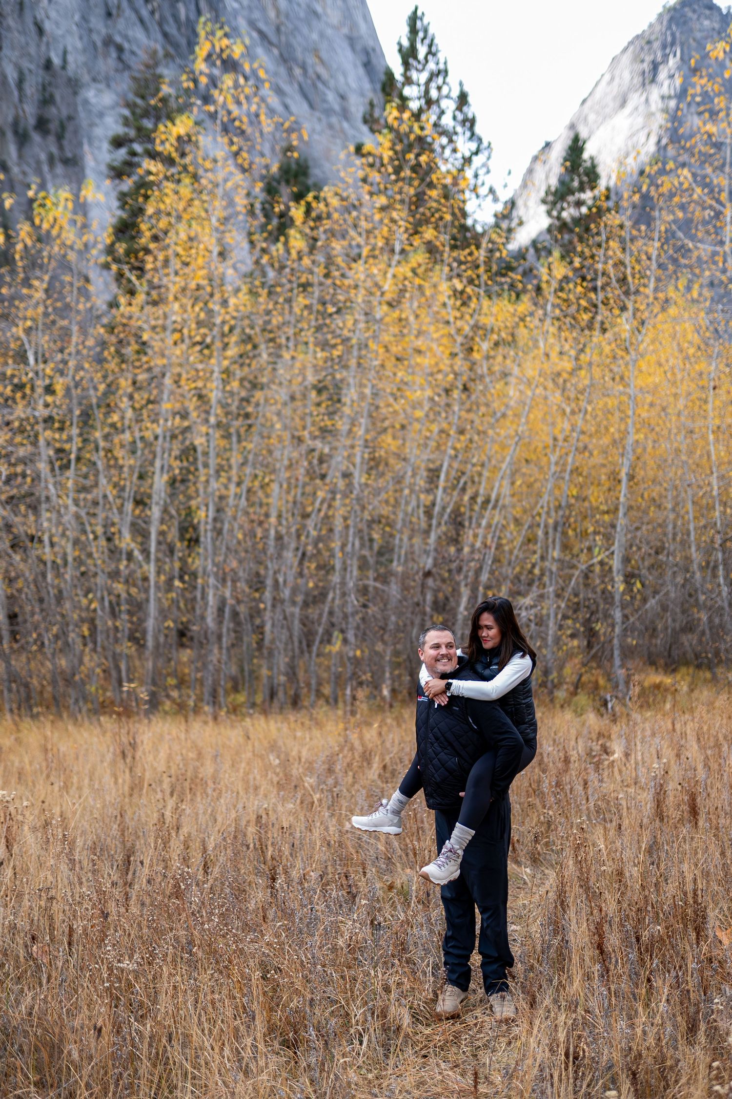 A couple embraces playfully in a field of tall grass surrounded by golden autumn trees and mountain scenery.